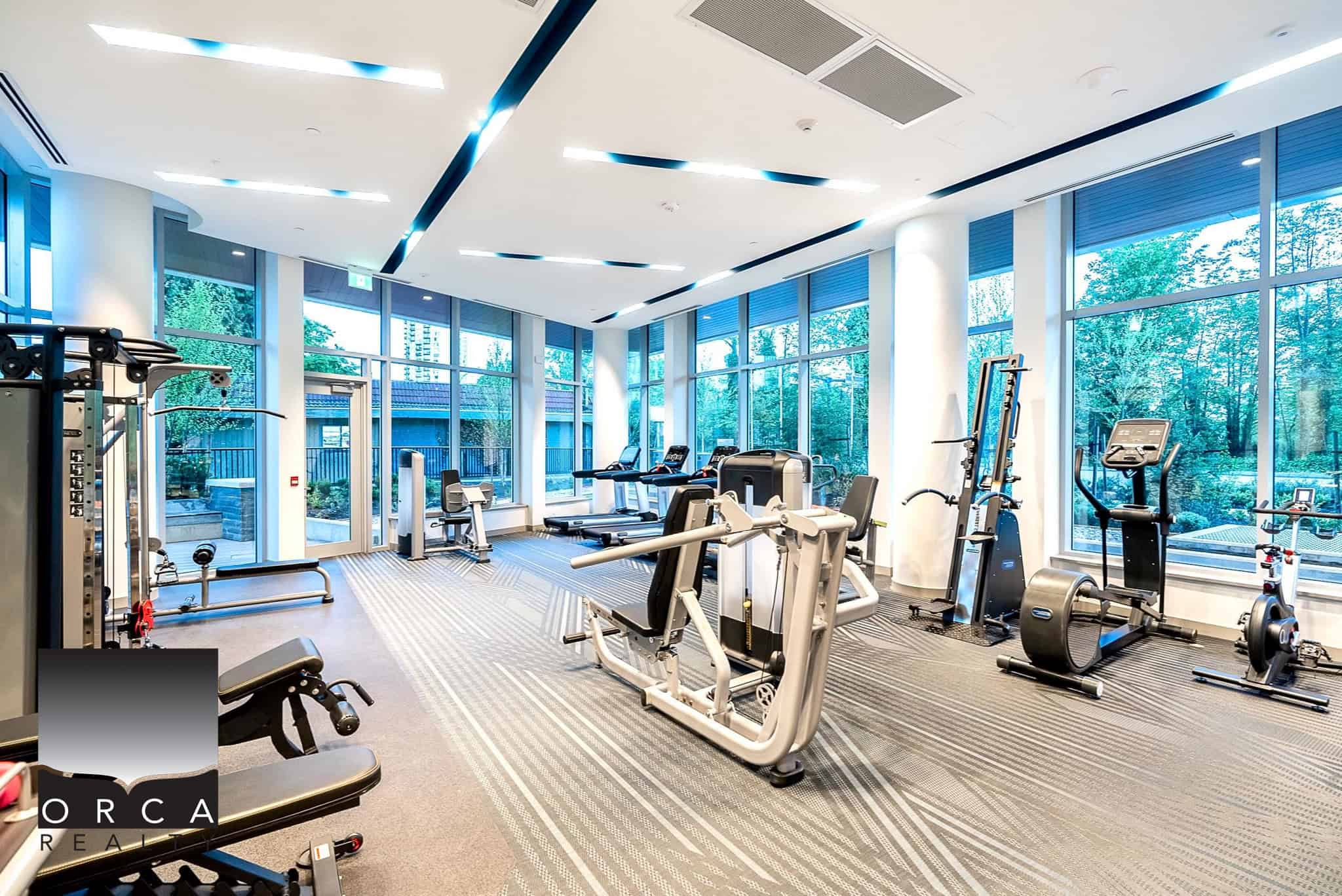 Modern fitness centre with cardio and strength training equipment, large windows, and natural light at Orca Realty Inc. offering premium real estate services in BC.