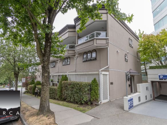 Modern multi-family residential building with arched windows, balconies, and beige exterior in an urban setting, surrounded by lush trees and well-maintained landscaping.