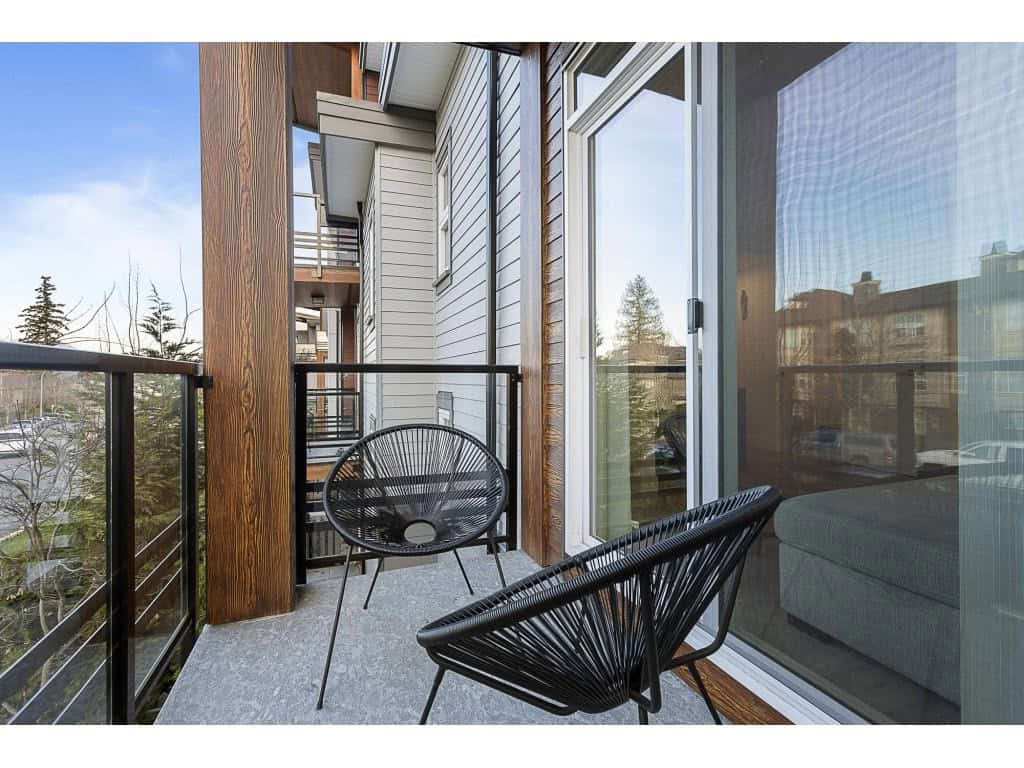 Modern apartment balcony with black outdoor chairs, wooden accents, and glass sliding door in a contemporary residential building. Perfect for outdoor relaxing and enjoying city or neighborhood views.