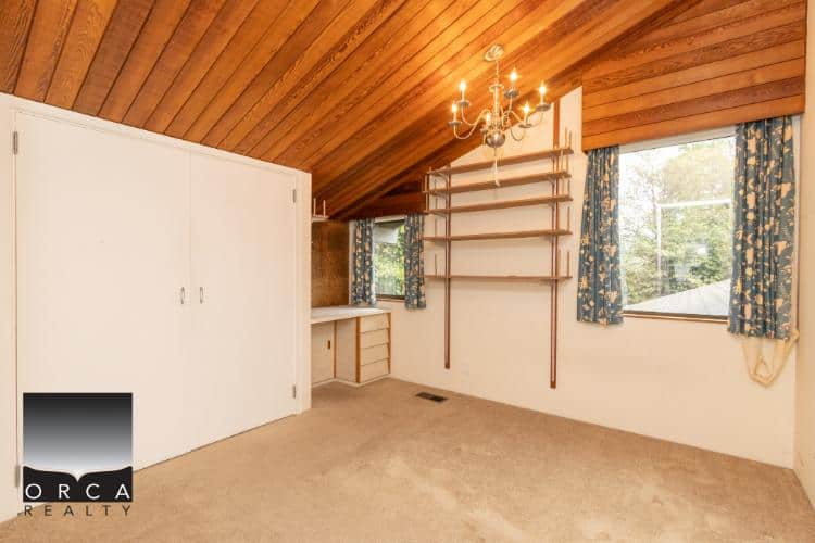 Bright interior room with sloped wooden ceiling, built-in storage, and large windows with floral curtains, showcasing a cozy and spacious area in a Vancouver Island home listed by Orca Realty Inc.
