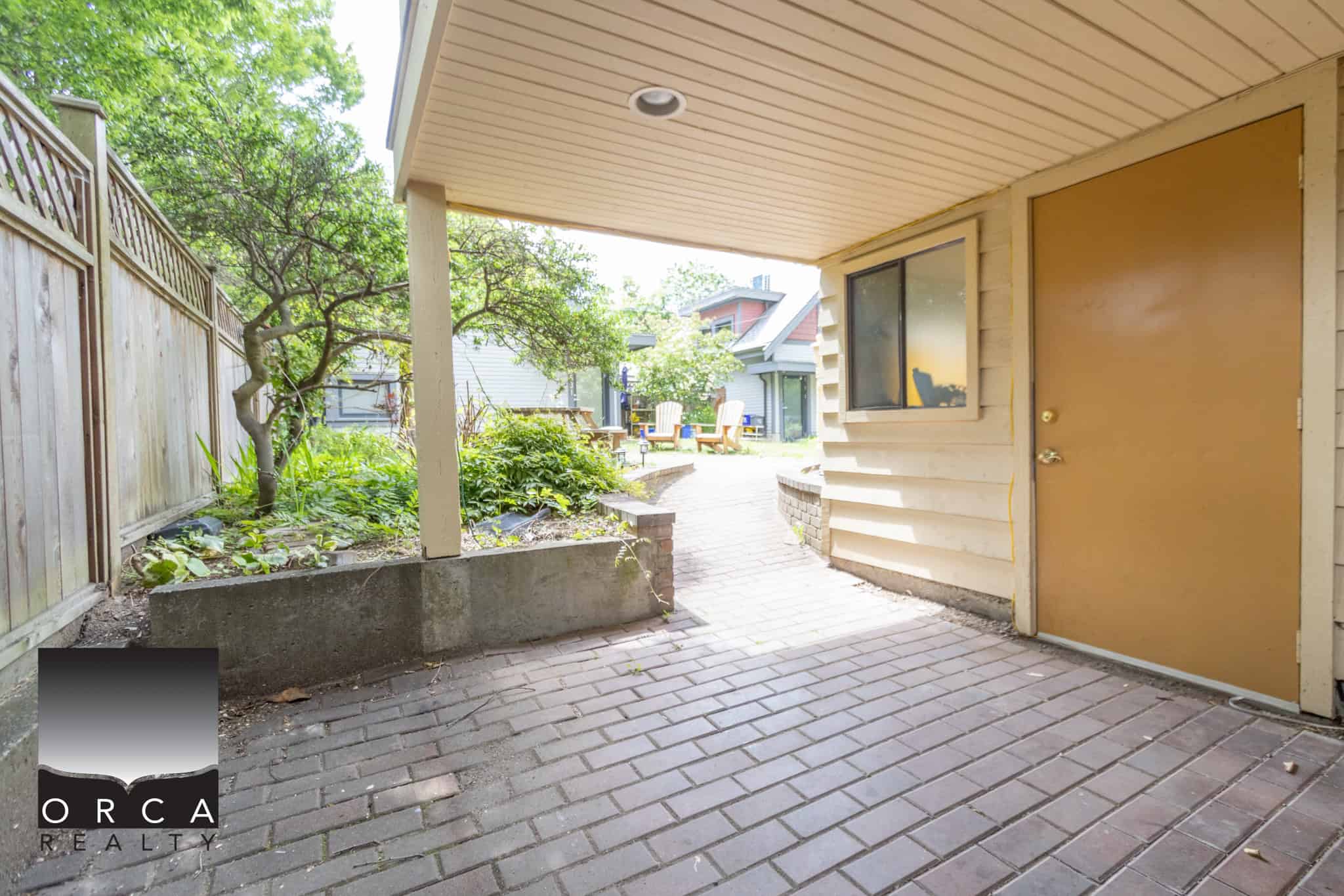 Outdoor patio area with brick flooring, covered porch, garden space with greenery, and backyard seating, perfect for relaxing or entertaining in a residential property listed by Orca Realty Inc.