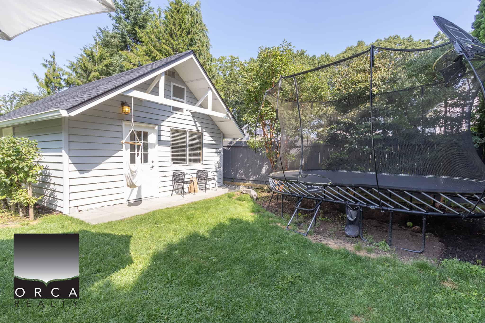 31 Peaceful backyard with a cozy grey cottage, green lawn, and a large outdoor trampoline, perfect for family fun and outdoor living in Vancouver Island.