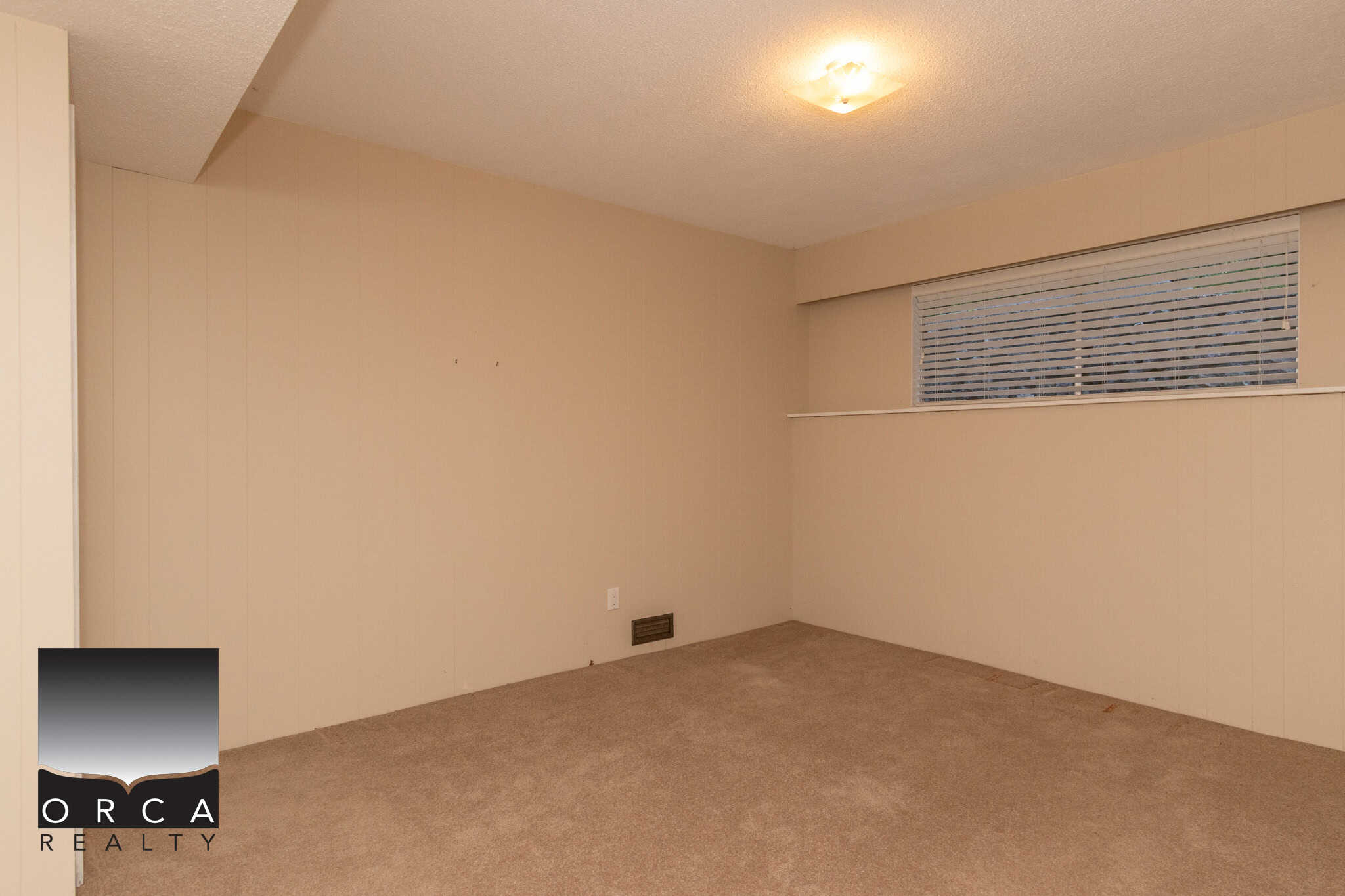 Spacious beige-carpeted living room with beige walls, a long horizontal window with white blinds, and a ceiling light fixture, ideal for Vancouver area homes.