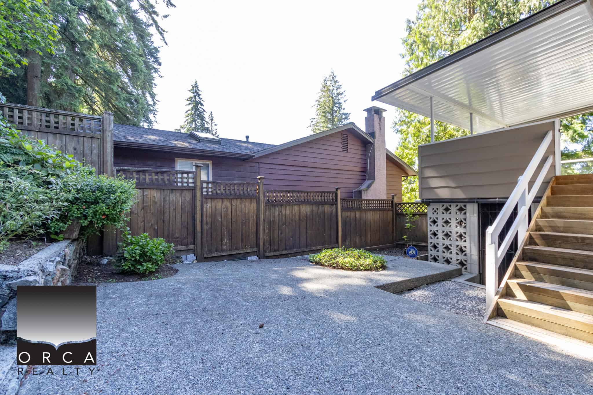 Spacious backyard with wooden fence, concrete patio, and wooden staircase leading to the house, surrounded by mature trees and lush greenery, ideal for outdoor living and entertaining.