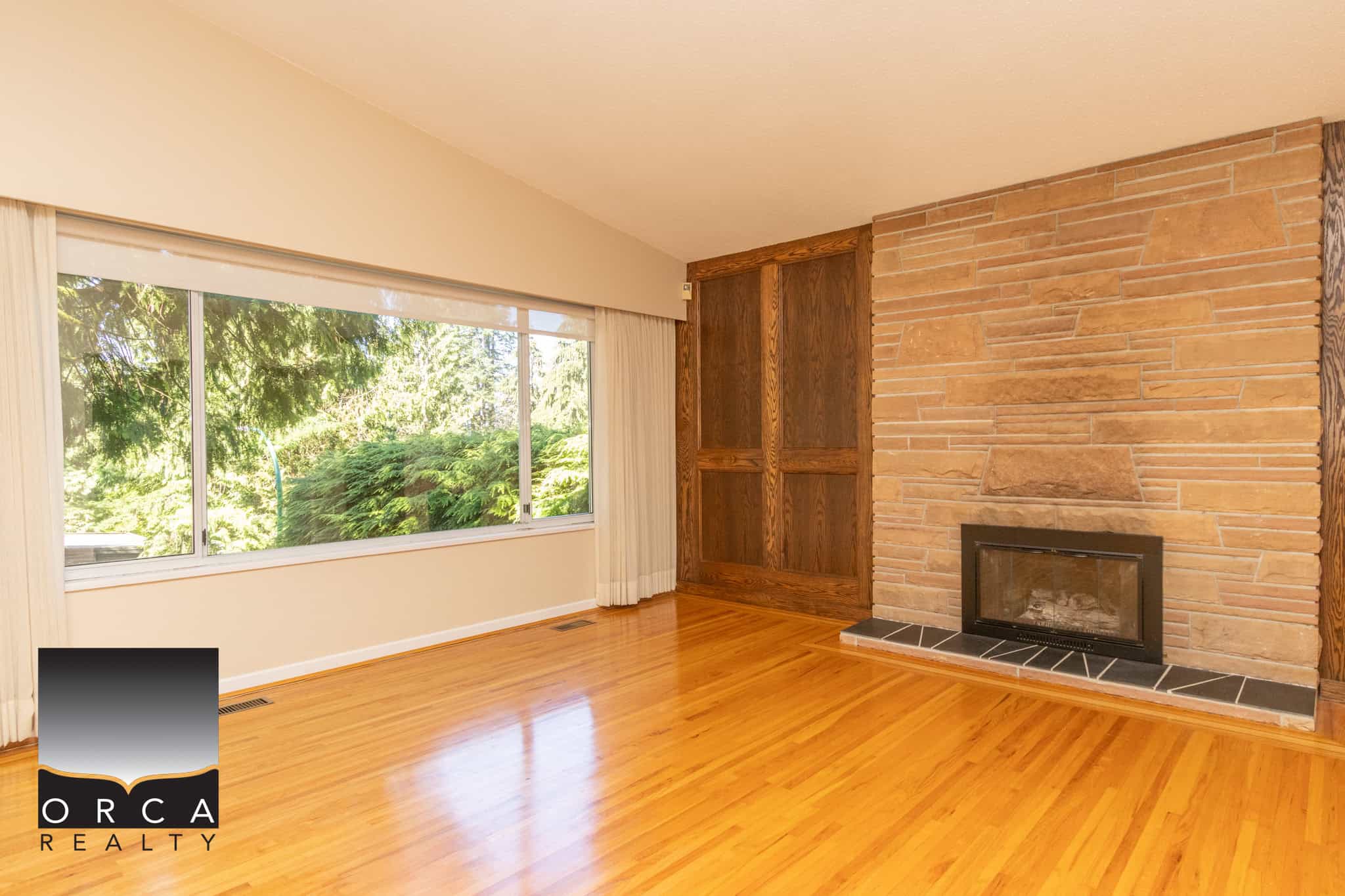 Spacious living room with large window, hardwood floors, and a cozy brick fireplace, ideal for warm gatherings and family space in a Vancouver area home listed by Orca Realty Inc.