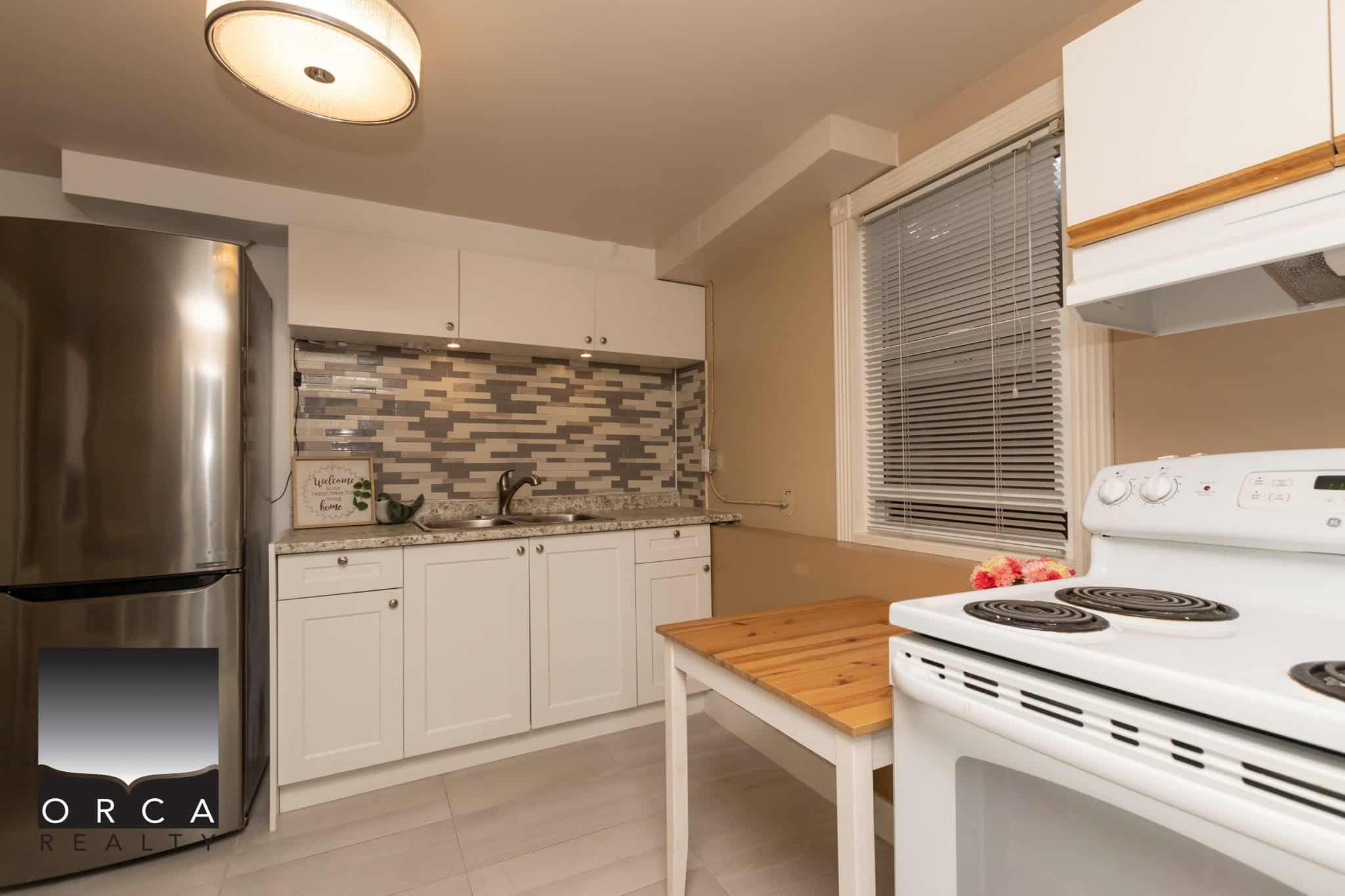 Stainless steel refrigerator, white kitchen cabinets, and a white stove in a modern kitchen with a mosaic tile backsplash. The space features a wooden countertop and a window with blinds, ideal for Vancouver property listings.