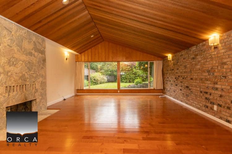 Spacious living room with wood-paneled ceiling, brick accent wall, and large window showcasing a lush backyard. Perfect for family gatherings or relaxing in a charming, sunny space.