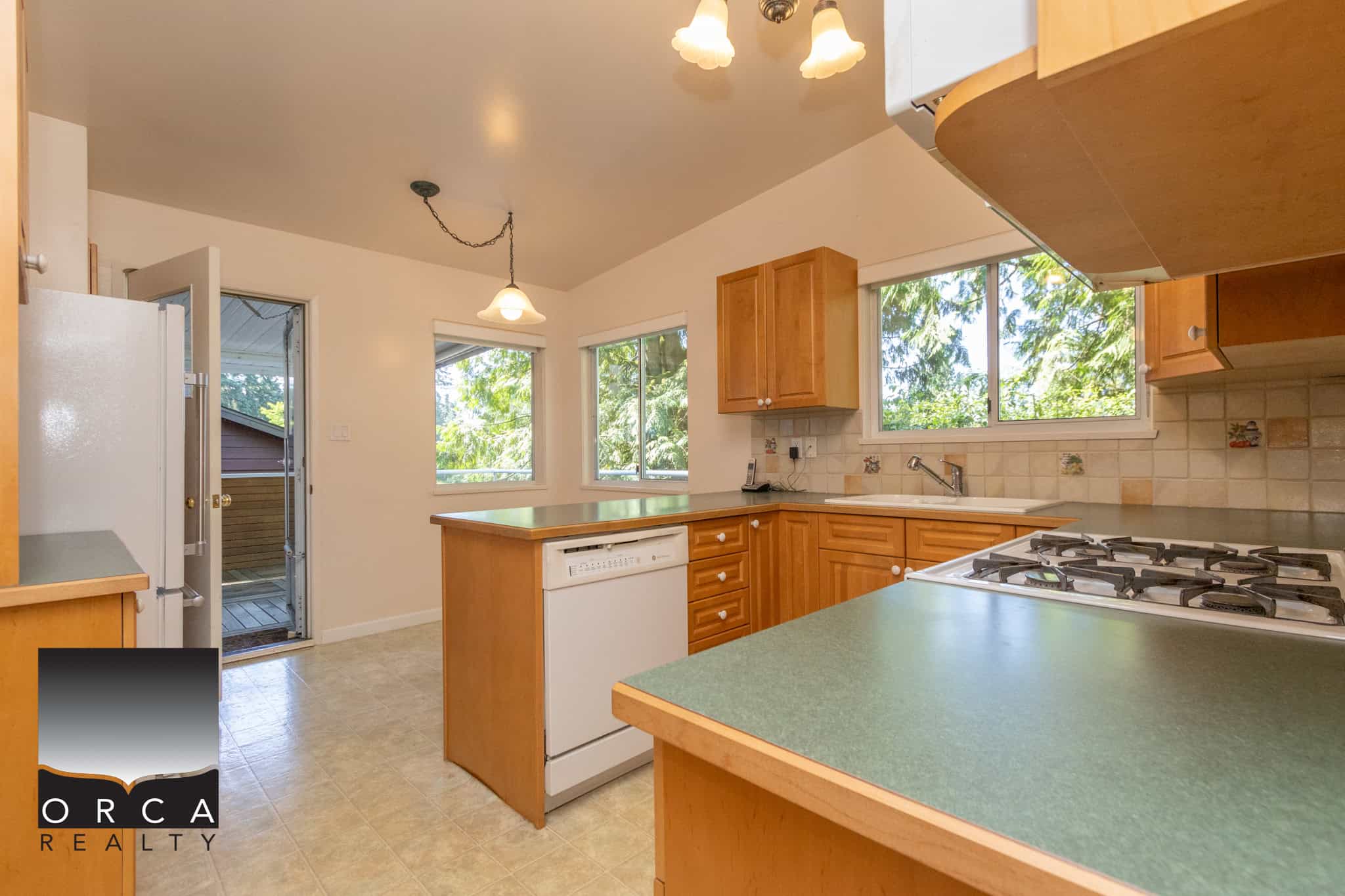 Bright kitchen with natural light, wooden cabinets, and green countertops, featuring a view of lush trees through the windows, perfect for family living and seamless indoor-outdoor access.