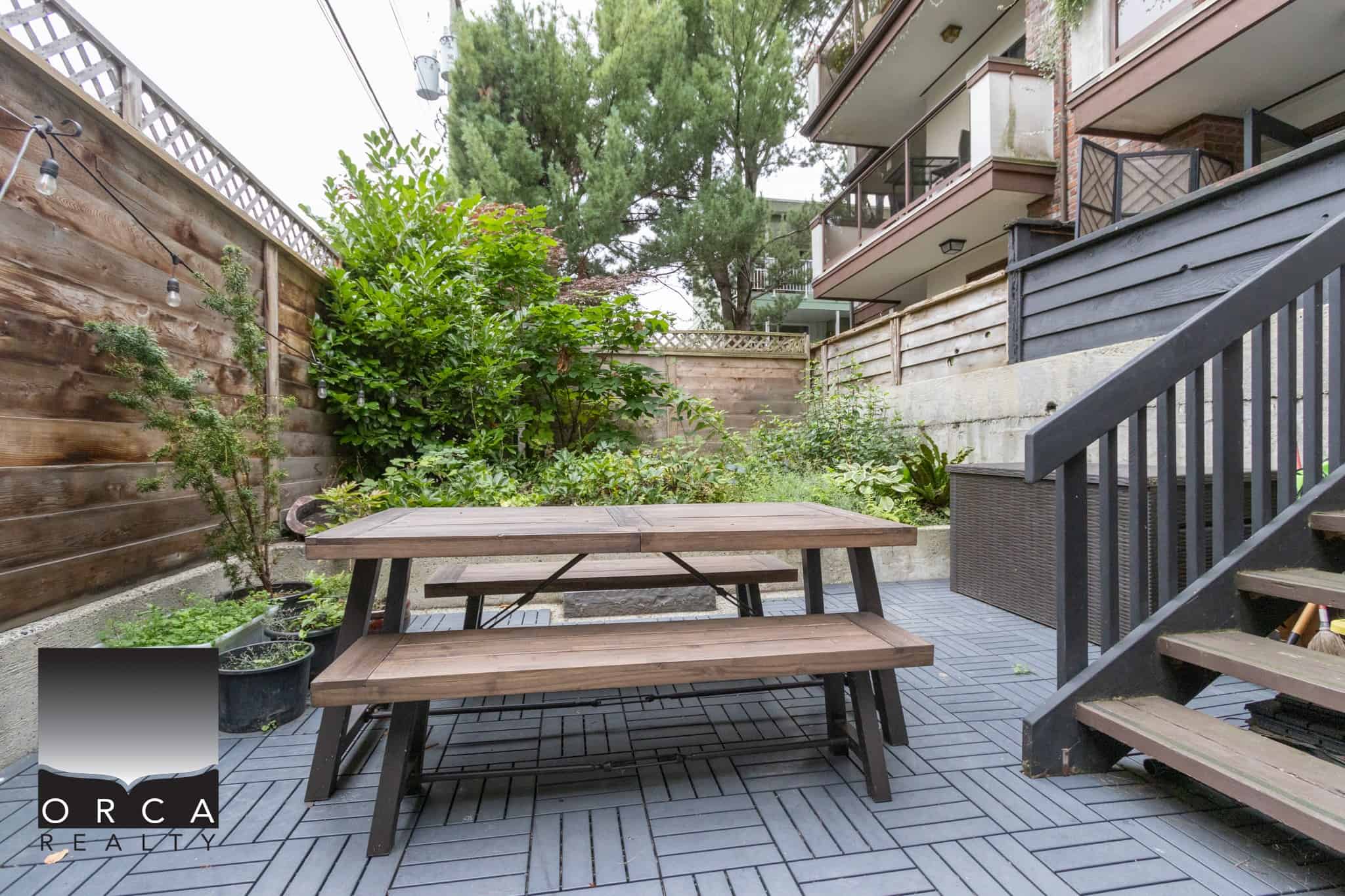 Cozy urban backyard patio with wooden picnic table, lush greenery, modern black staircase, enclosed by wooden fencing, perfect for outdoor dining and relaxation in Vancouver.
