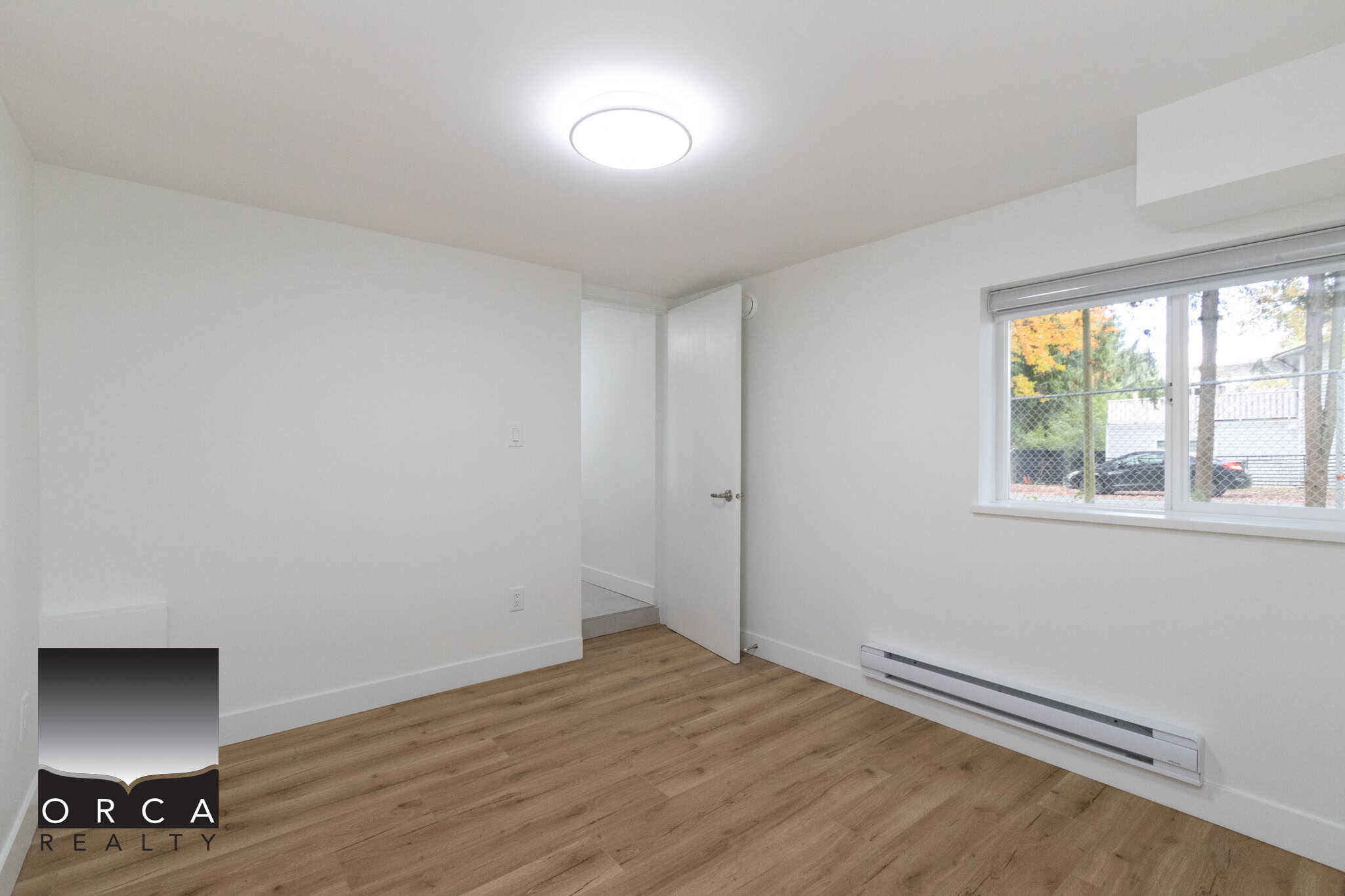 Bright empty room with white walls, wooden flooring, and a large window in a modern home listing by Orca Realty Inc.