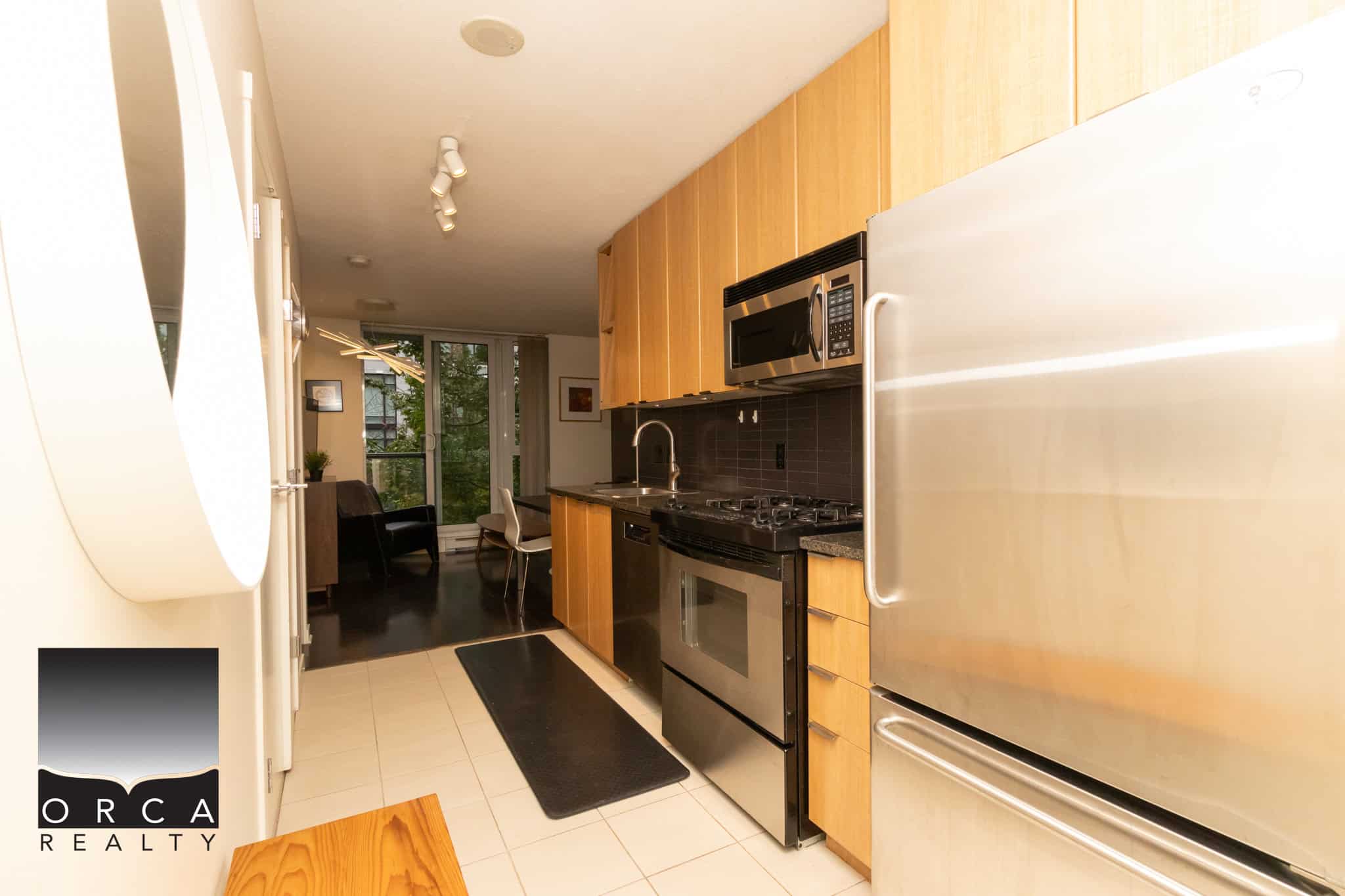Modern Vancouver apartment kitchen with stainless steel appliances and wooden cabinets, open to a dining and living area featuring a balcony with outdoor greenery.