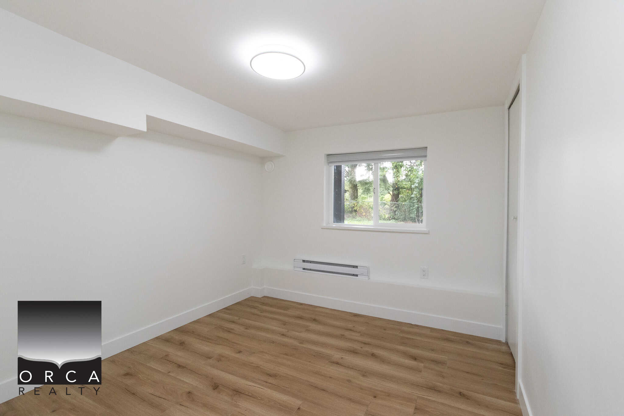 Bright empty room with white walls, wooden flooring, and a window overlooking a green outdoor area, ideal for home staging or real estate listings.