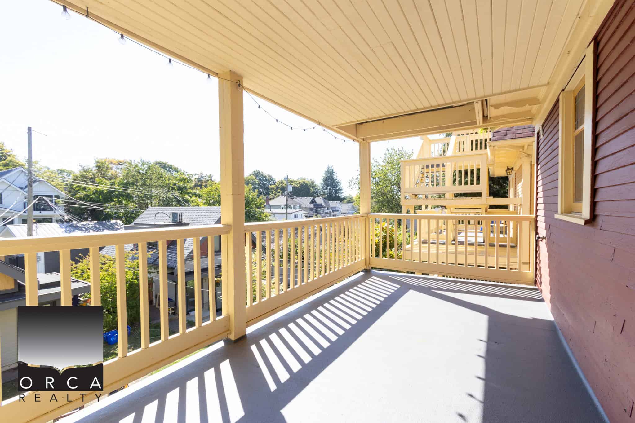 Spacious residential balcony with wooden railing and overhead string lights, overlooking a neighbourhood with trees and houses, perfect for outdoor relaxation and entertaining.