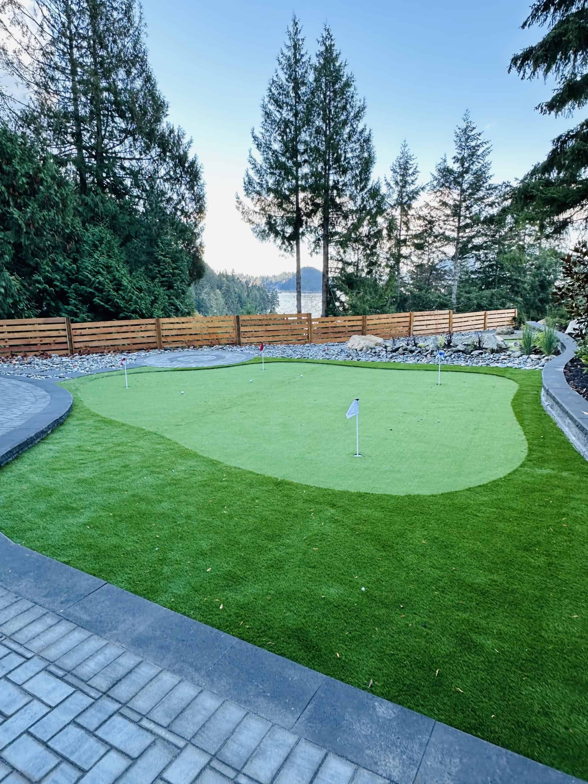 Lush backyard putting green with surrounding stone pathway, tall evergreen trees, and a wooden privacy fence, perfect for outdoor recreation and enjoying nature in Vancouver Island.