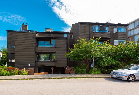 Contemporary multi-unit residential building with dark exterior siding in a Vancouver neighbourhood, featuring balconies, lush greenery, and parking spaces.
