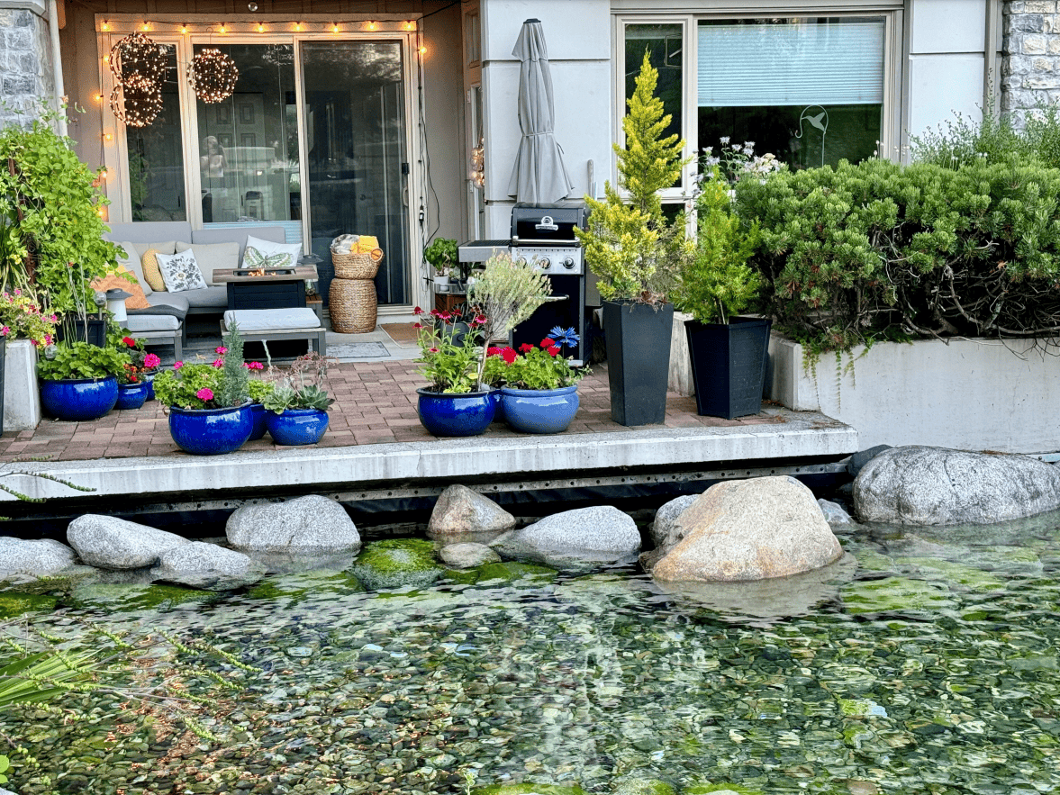 Flower pots and outdoor patio with seating and barbecue on waterfront property, lush greenery, cozy backyard, decorative lighting, outdoor living space, garden, landscape design, real estate, Vancouver Island, property listing.