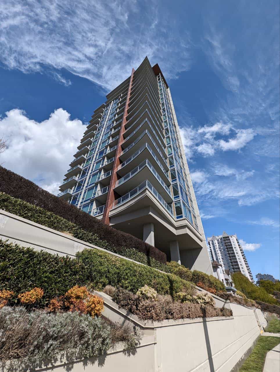 Modern high-rise residential building with glass balconies, located in a vibrant urban area with landscaped surroundings and clear blue sky, representing luxury real estate in Vancouver.