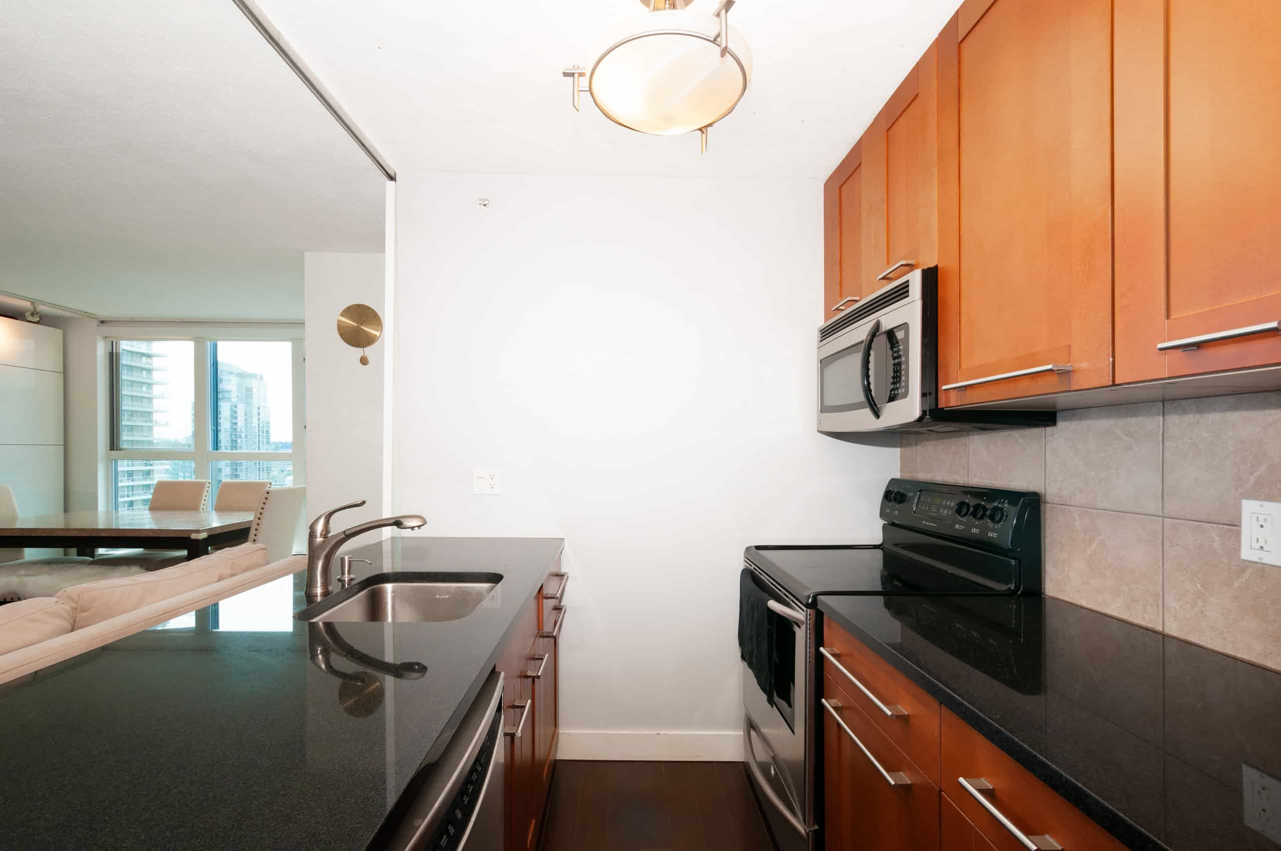 016 Contemporary kitchen in Vancouver condo with cherry wood cabinets, black granite countertops, stainless steel appliances, and open concept living space.
