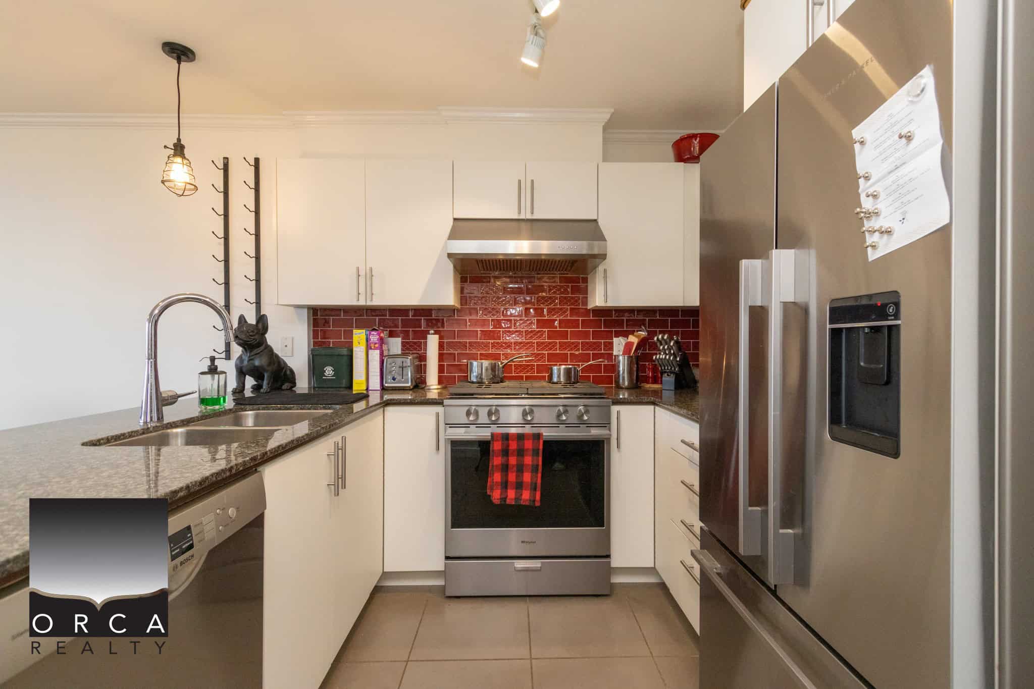 Modern kitchen with stainless steel appliances, white cabinetry, red brick backsplash, and granite countertops in a contemporary Orca Realty Inc. listing.
