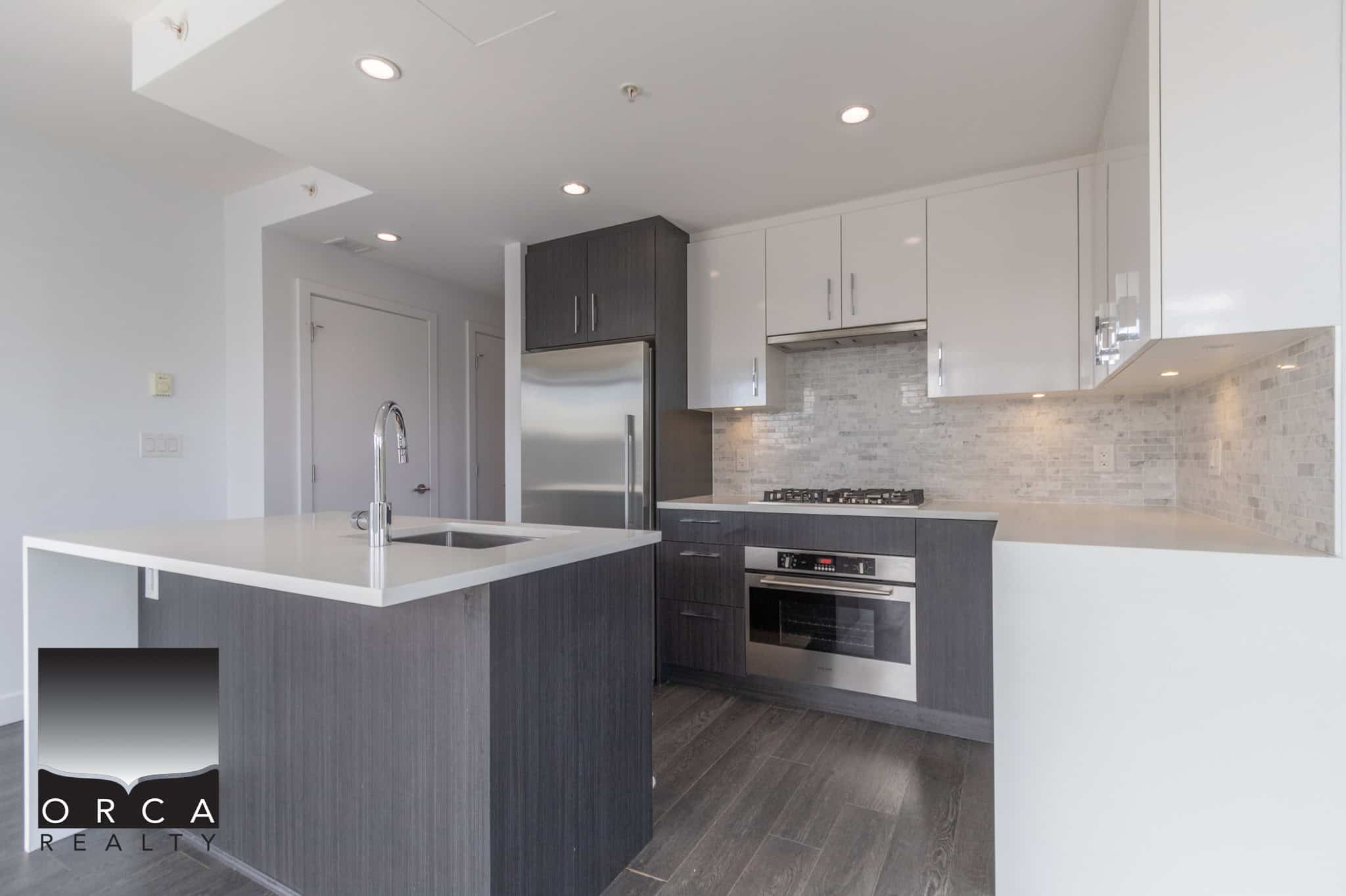 Modern kitchen with dark wood cabinetry, stainless steel appliances, and a sleek white island in a contemporary Orca Realty Inc. listed property.