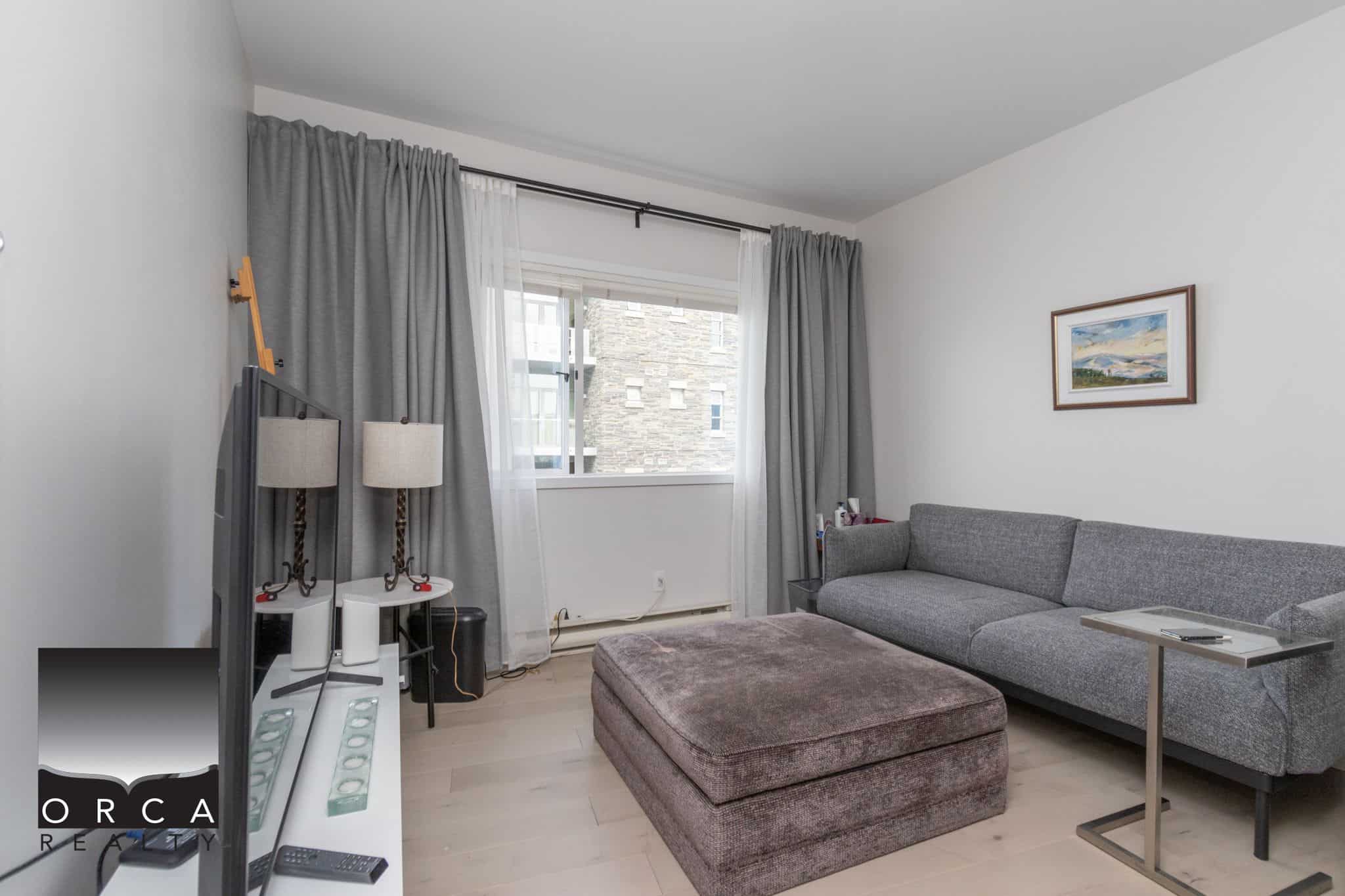 Cozy living room with a large gray sofa, ottoman, and modern decor in a Vancouver property. Bright space featuring a window with curtains and simple furnishings, ideal for comfortable urban living.
