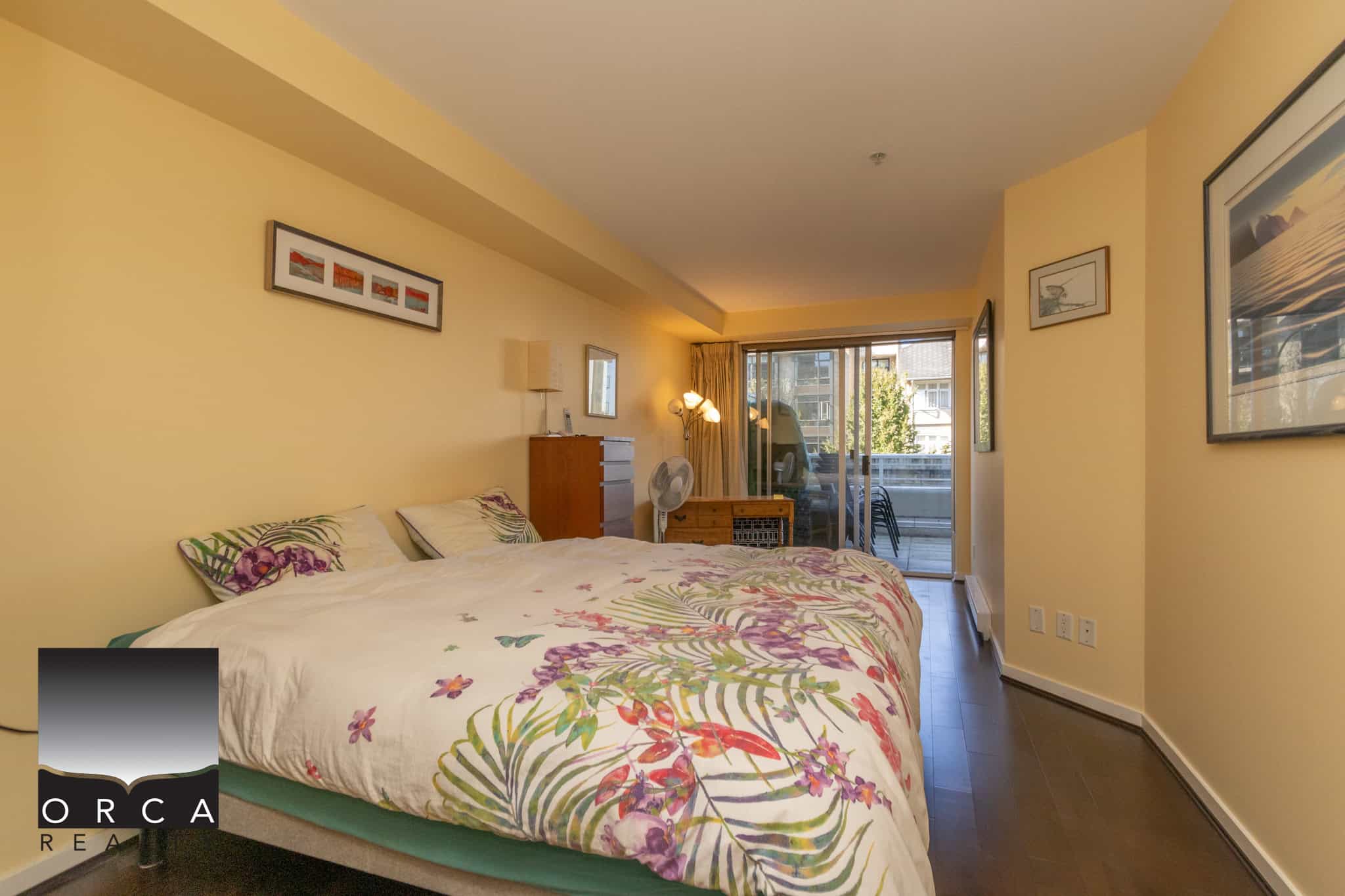 Bright bedroom with sliding glass door leading to balcony, featuring soft lighting, framed art, and cozy bedding, ideal for Vancouver Coastal real estate listings.