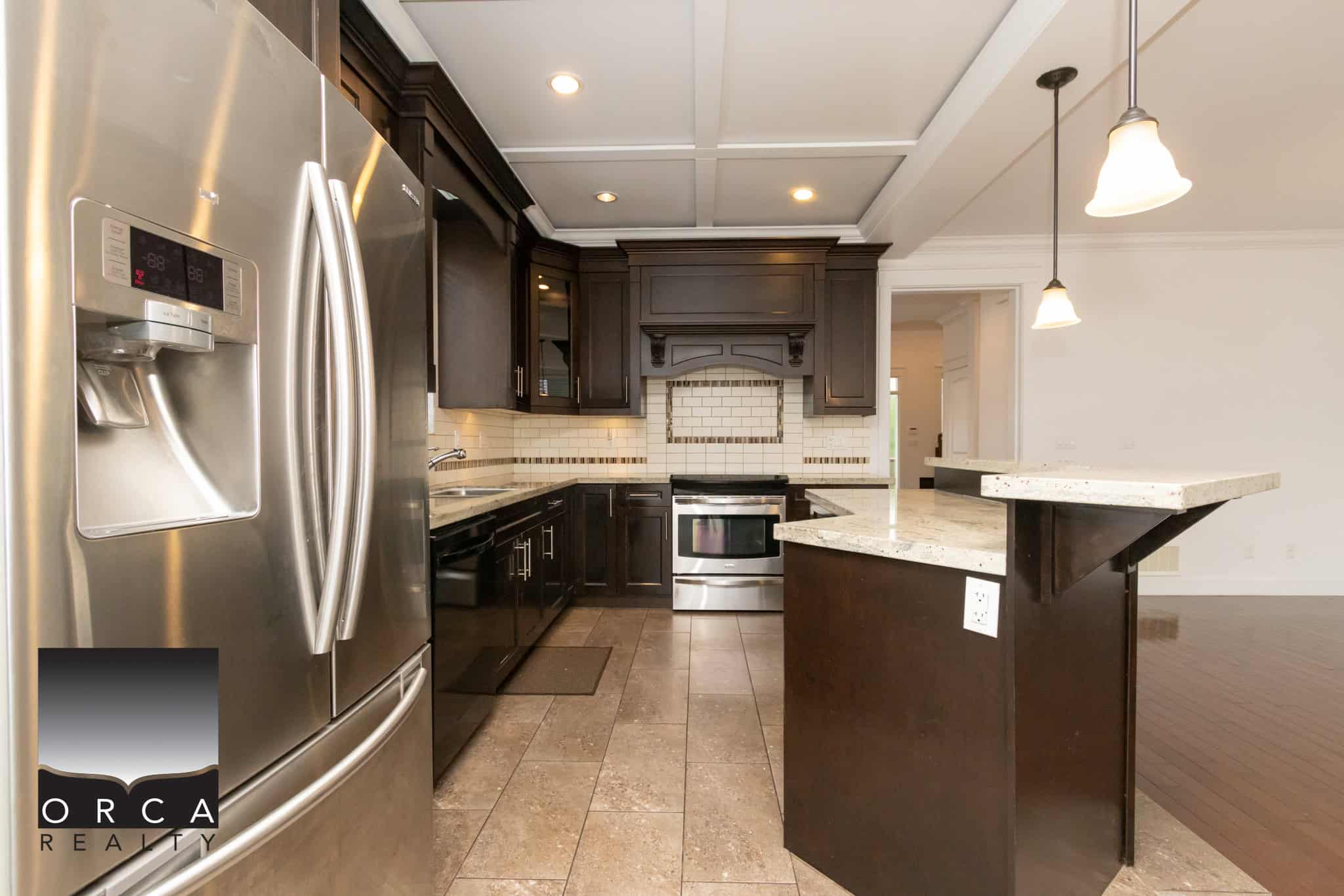 Stainless steel refrigerator in a modern kitchen with dark cabinetry, granite countertops, and contemporary pendant lighting, showcasing a luxurious home interior by Orca Realty Inc.