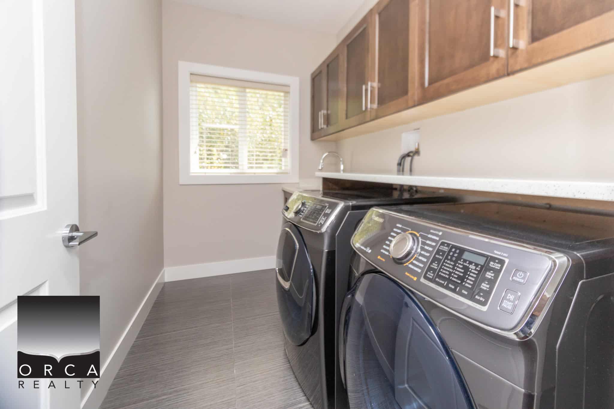 Modern laundry room with front-load washer and dryer, wooden cabinetry, neutral wall color, and natural light from a window, ideal for home comfort and convenience.