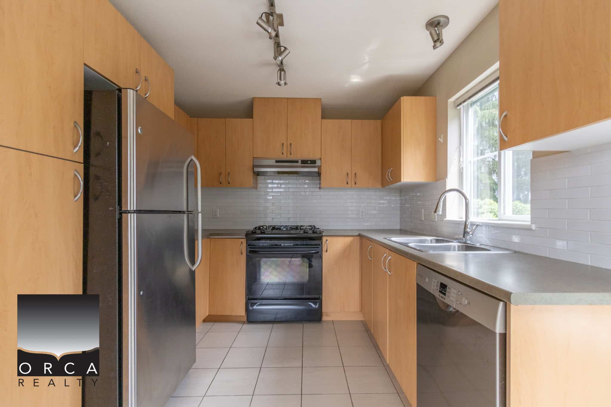 Stainless steel refrigerator in a modern kitchen with wooden cabinetry, white tile backsplash, and a double stainless steel sink, showcasing a clean and functional Vancouver home interior.