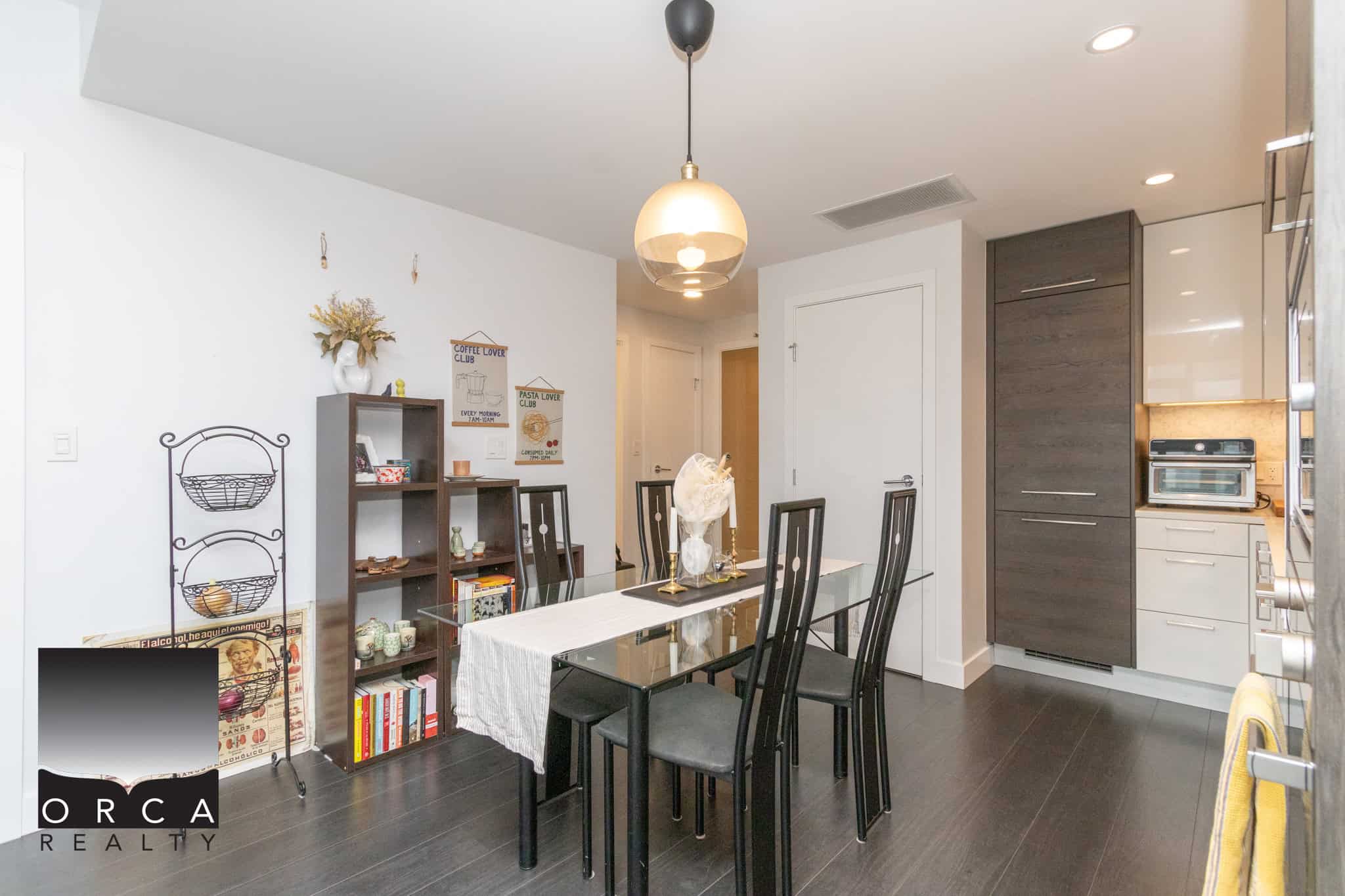 Modern dining room with dark wood flooring, glass dining table, black chairs, and contemporary kitchen cabinetry, featuring a cozy and stylish decor ideal for Vancouver real estate listings.