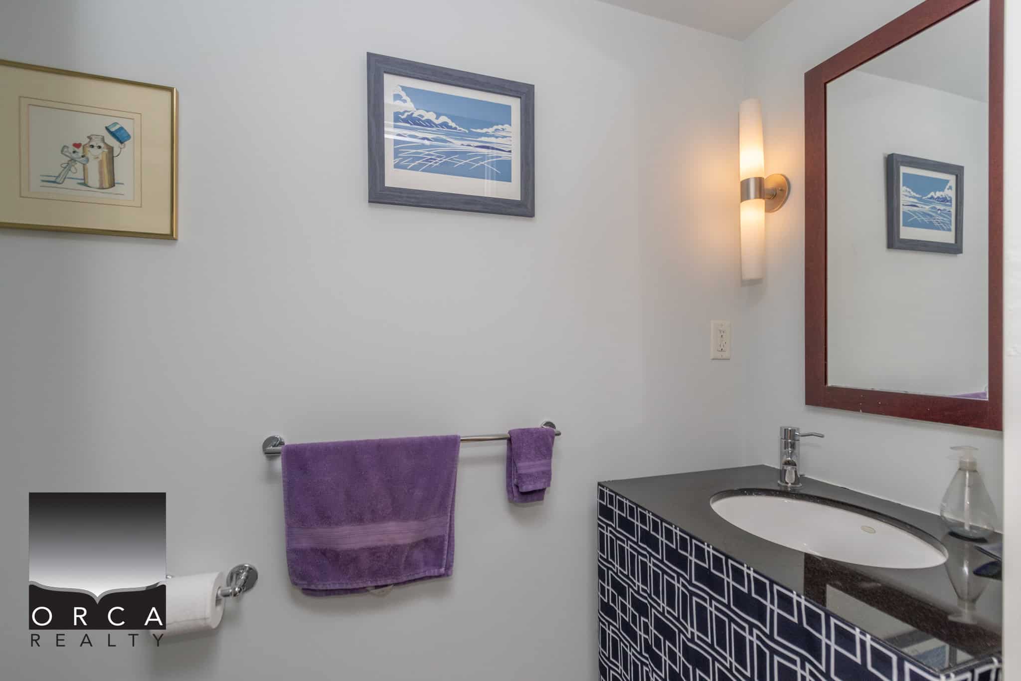 Vancouver Island real estate bathroom with modern decor, purple towels, framed ocean art, and a sleek vanity with mirror, reflecting Orca Realty Inc. branding for optimal SEO.
