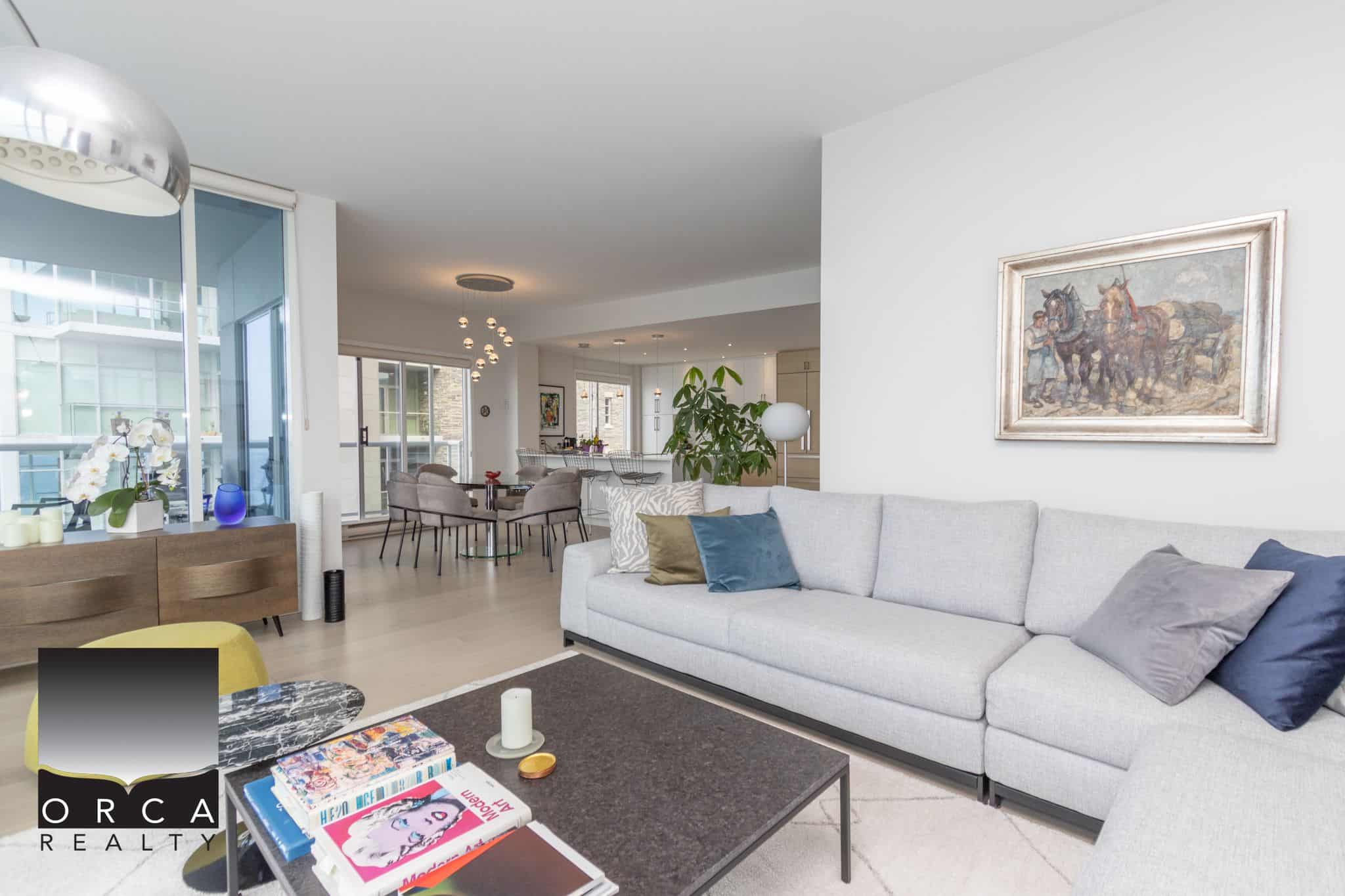 Bright modern living room interior with white sofa, decorative cushions, artwork, and open concept kitchen and dining area, featuring natural light and contemporary decor.