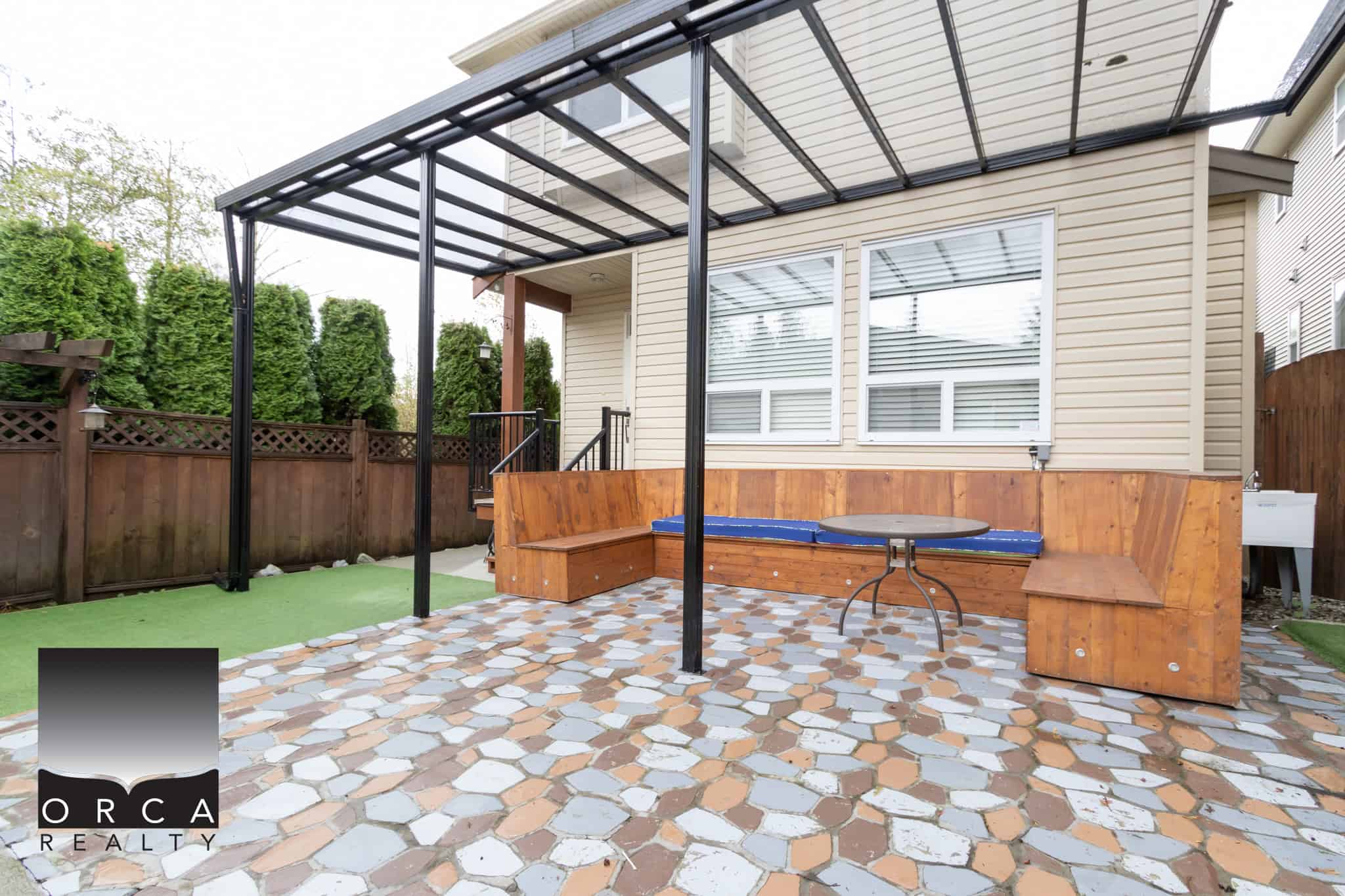 Colorful outdoor patio area with wooden bench seating, small round table, covered by a metal pergola, surrounded by a privacy fence and lush green trees, perfect for relaxing outdoors.