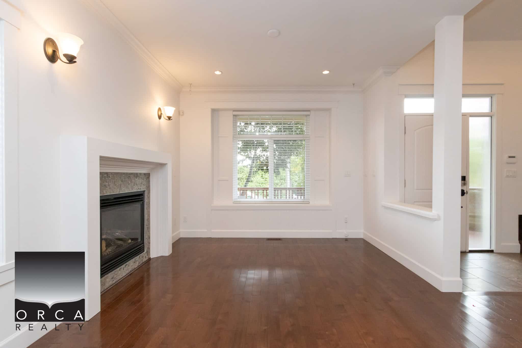 Bright living room with a fireplace, hardwood floors, large window, and stylish wall sconces, showcasing a welcoming space perfect for family and entertaining in a home listed by Orca Realty Inc.