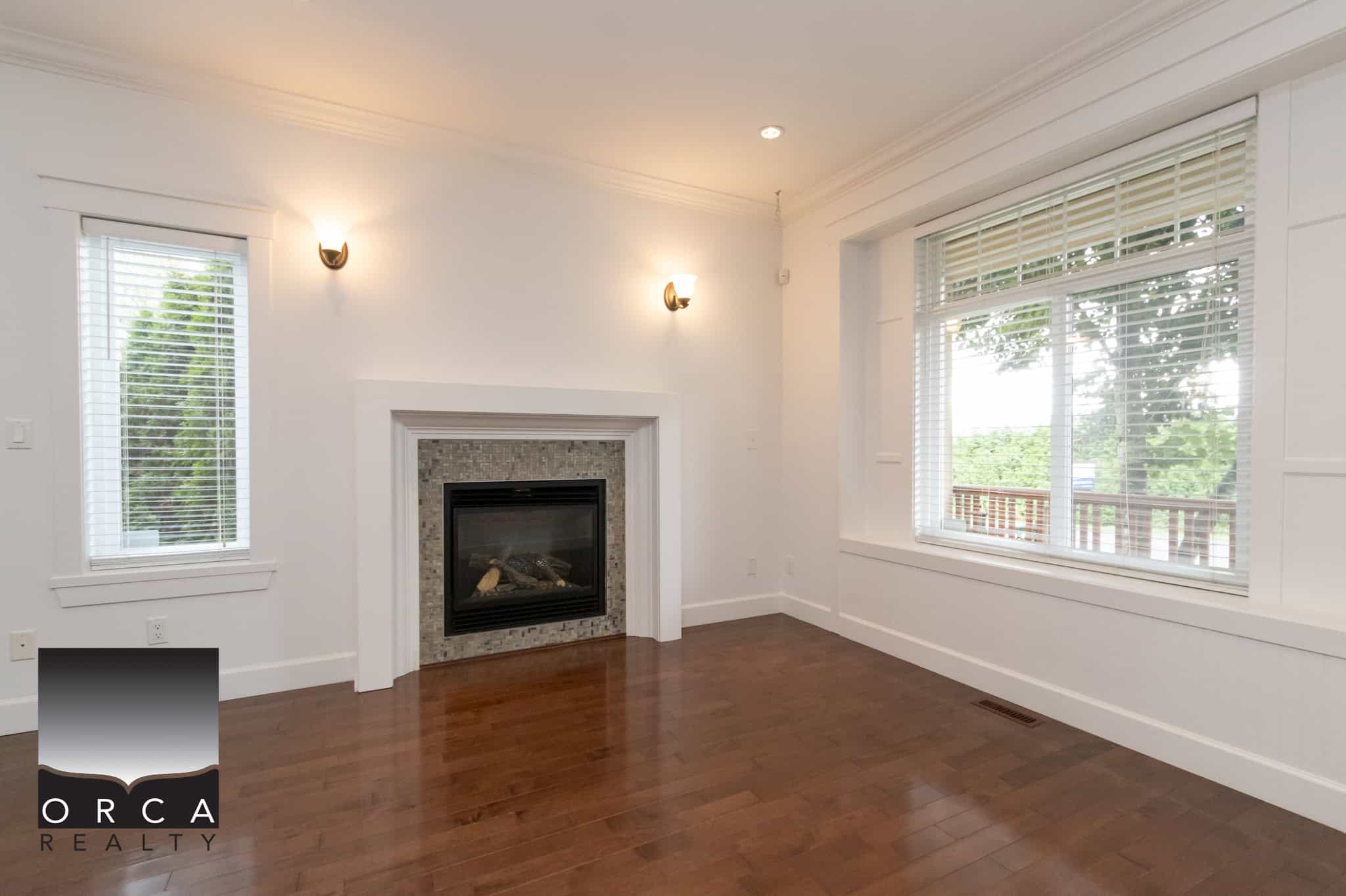 Bright living room with fireplace, large windows with white blinds, hardwood flooring, and modern lighting, showcasing a spacious and inviting interior in a property listed by Orca Realty Inc.