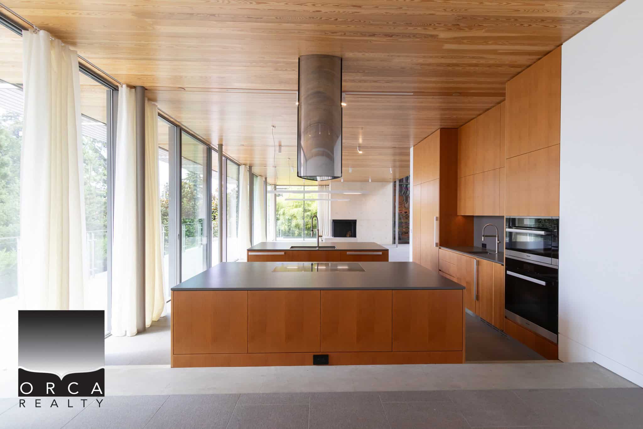 Modern open-concept kitchen with sleek wooden cabinetry, large island, and floor-to-ceiling windows showcasing natural light and greenery, perfect for contemporary home living.