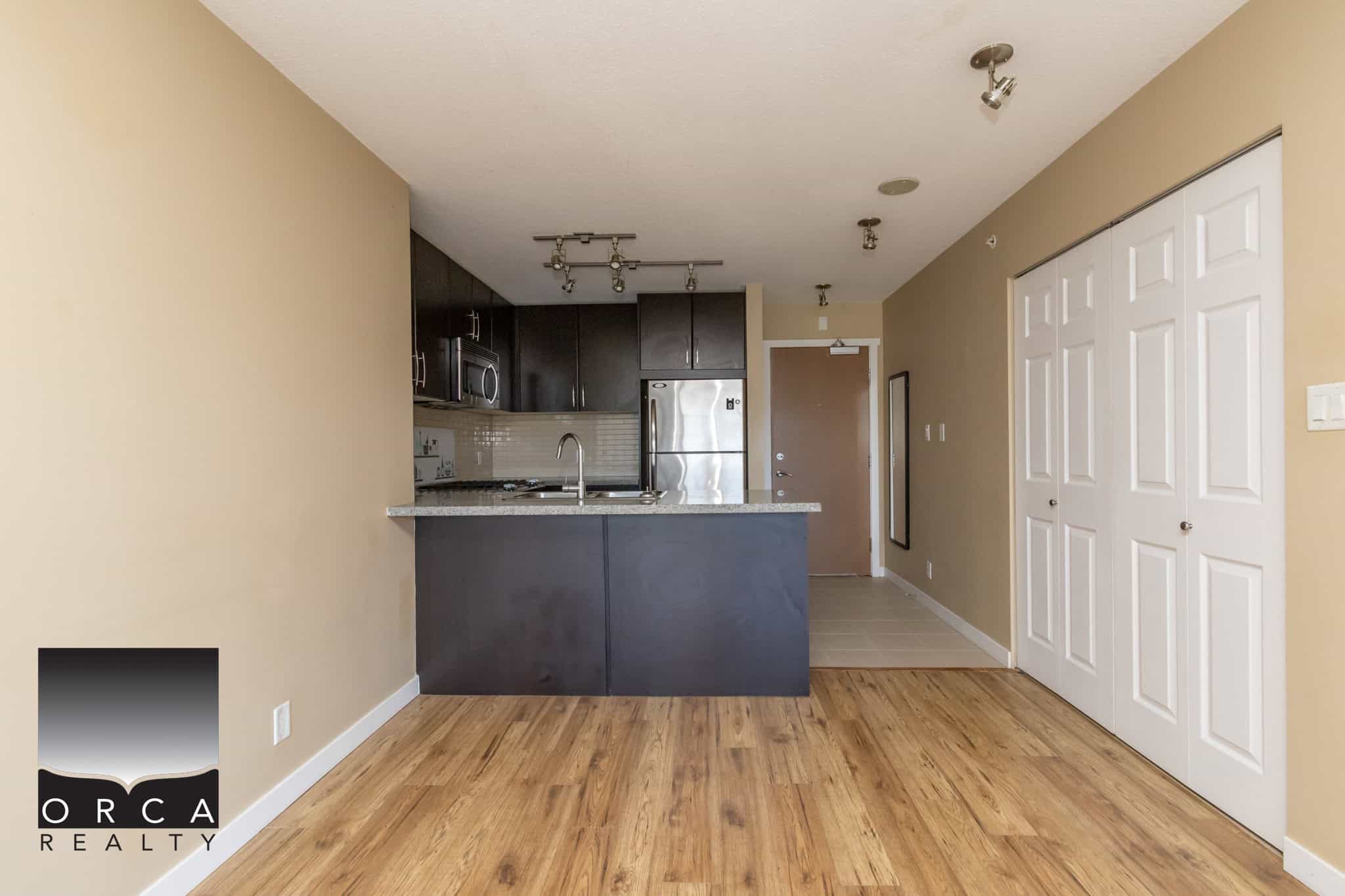 Modern kitchen and open living space in a Vancouver-area condo listed by Orca Realty Inc. featuring stainless steel appliances, granite countertops, and hardwood flooring.