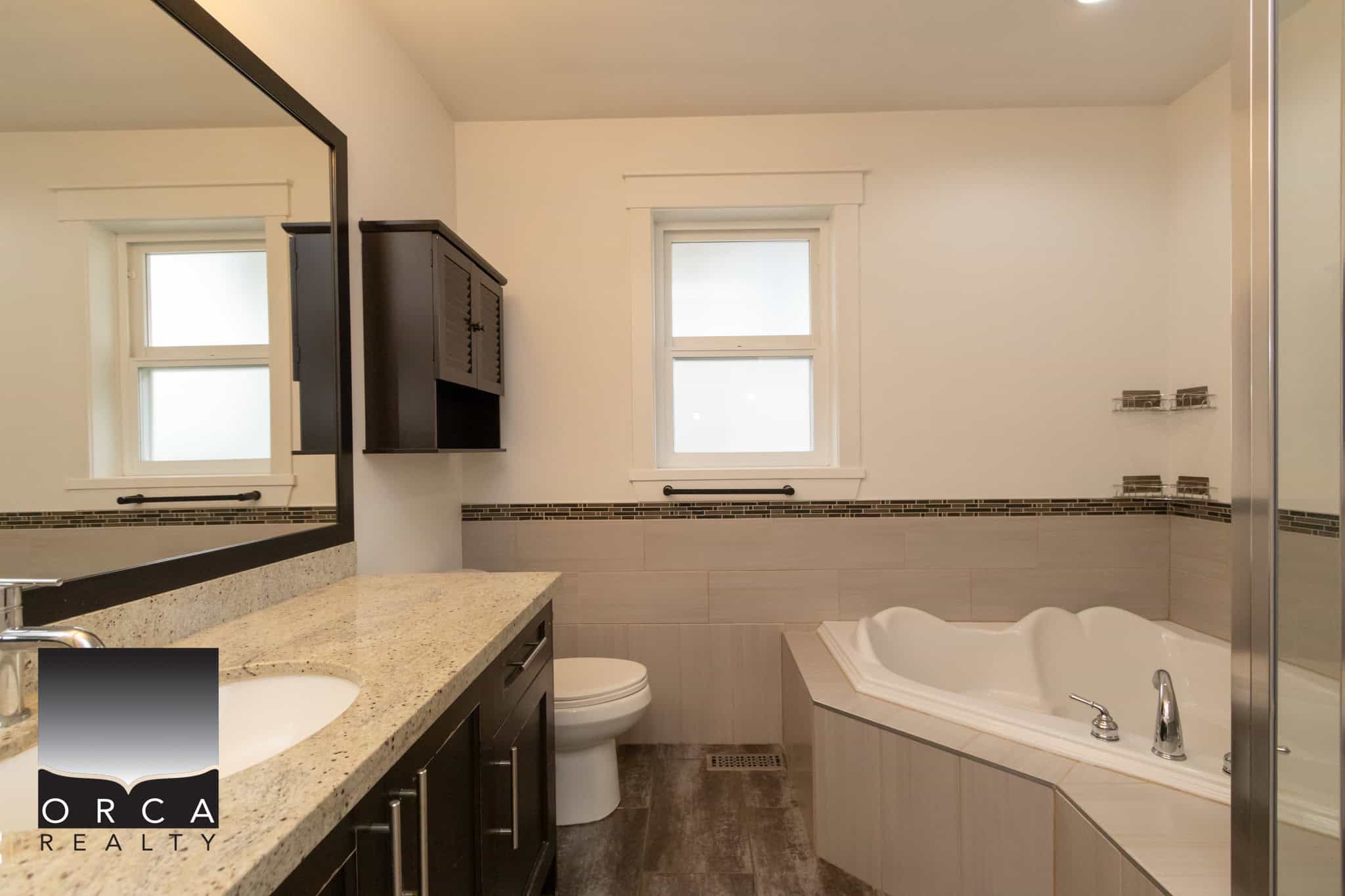 Bright bathroom with a double vanity, granite countertop, dark wood cabinets, large mirror, bathtub, and frosted windows, showcasing modern design and natural light in a home listed by Orca Realty Inc.