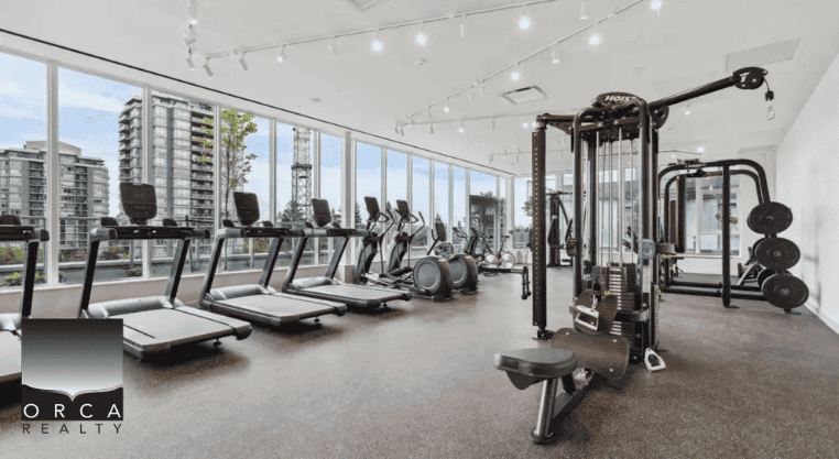 Modern gym interior at Orca Realty Inc. featuring cardio machines, weight equipment, and floor-to-ceiling windows with city views, ideal for active lifestyles in Vancouver real estate.