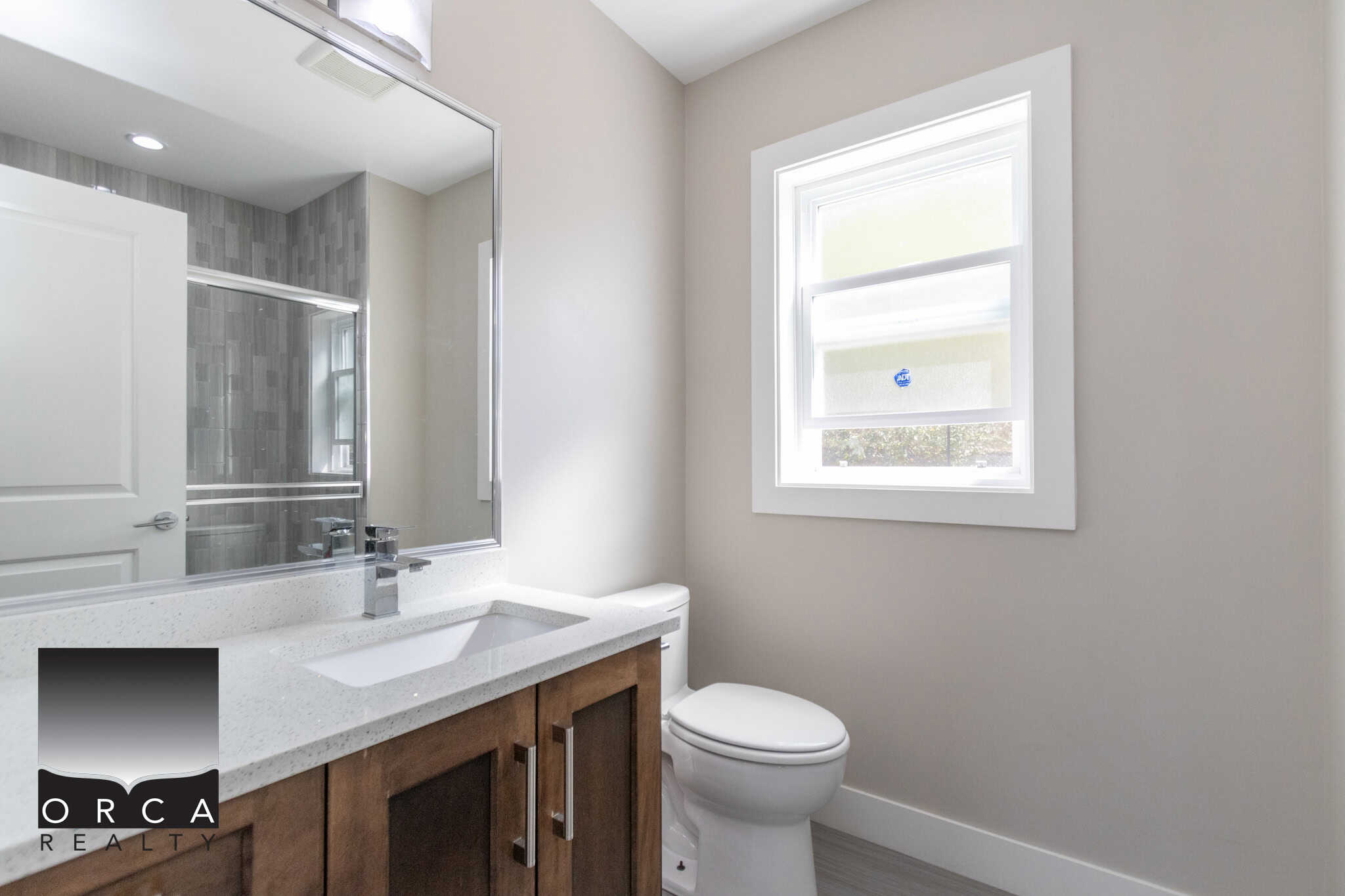 Modern bathroom with white walls, a sleek vanity with wooden cabinets, a large mirror, and a window providing natural light, showcasing contemporary real estate in the Orca Realty portfolio.