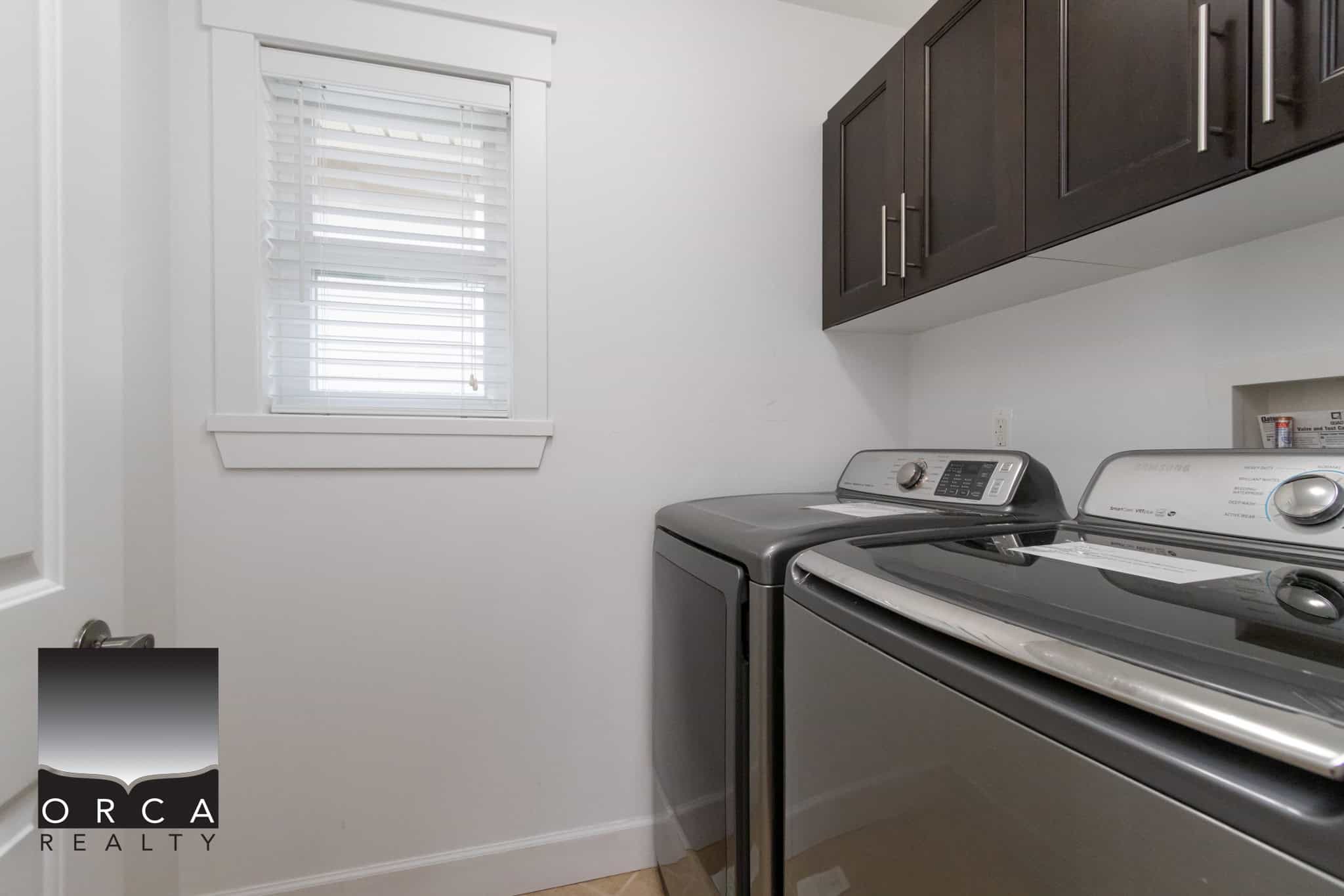 Modern laundry room with top-loading washer and dryer, white walls, dark wood cabinets, and a window with white blinds, ideal for convenient home living and property marketing.
