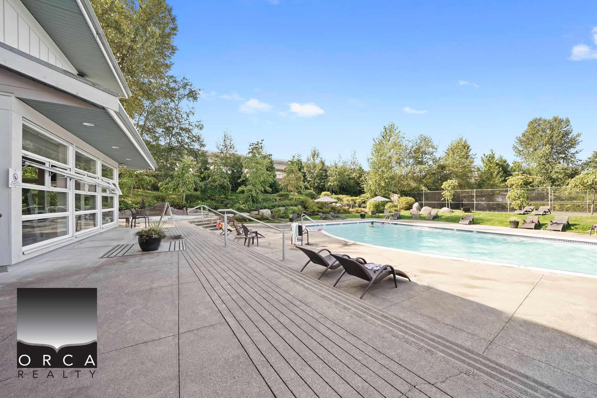 Luxury apartment building swimming pool area at Orca Realty Inc. showcasing outdoor seating with poolside lounge chairs, surrounded by lush greenery and modern amenities in a scenic setting.