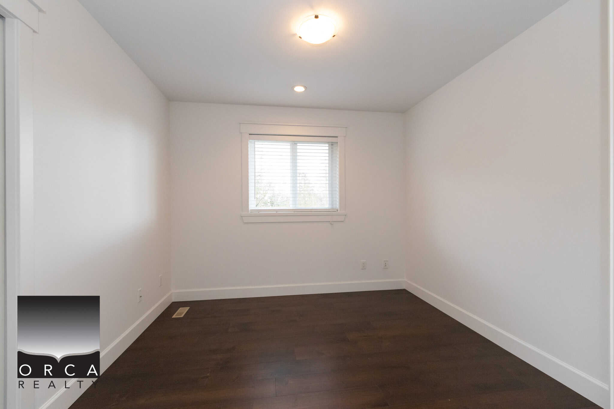 Bright empty bedroom with white walls and dark hardwood flooring, featuring a window with white blinds and a ceiling light, ideal for Vancouver real estate listings.