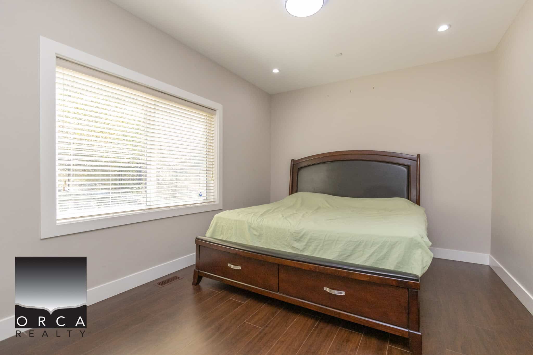 Bright and spacious bedroom in a modern home listed by Orca Realty Inc., featuring hardwood floors, a large window with blinds, and a stylish wooden bed frame with built-in storage, perfect for Vancouver Island homes.
