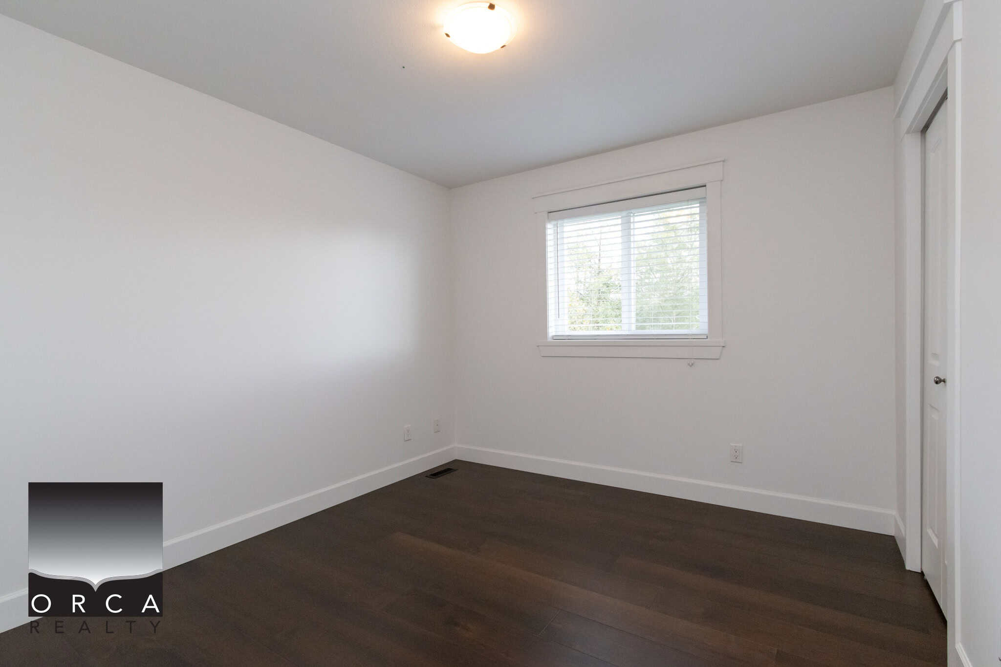 Bright empty bedroom with white walls, dark wood flooring, window with blinds, and closet door, ideal for Vancouver homebuyers seeking modern real estate in BC.