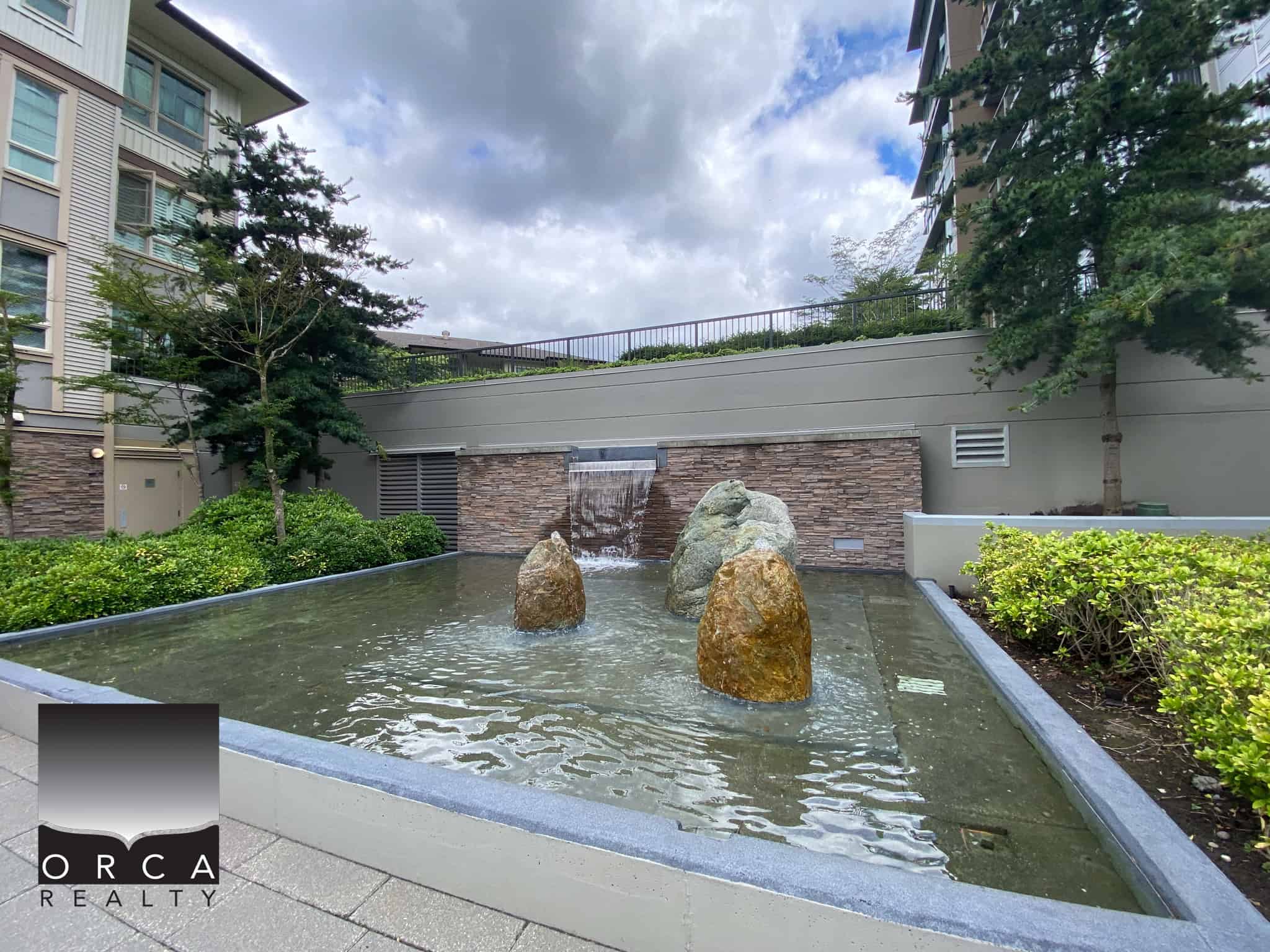 Modern outdoor water feature with rocks, lush greenery, and a cascading waterfall, located in a contemporary apartment complex courtyard in Vancouver, BC.
