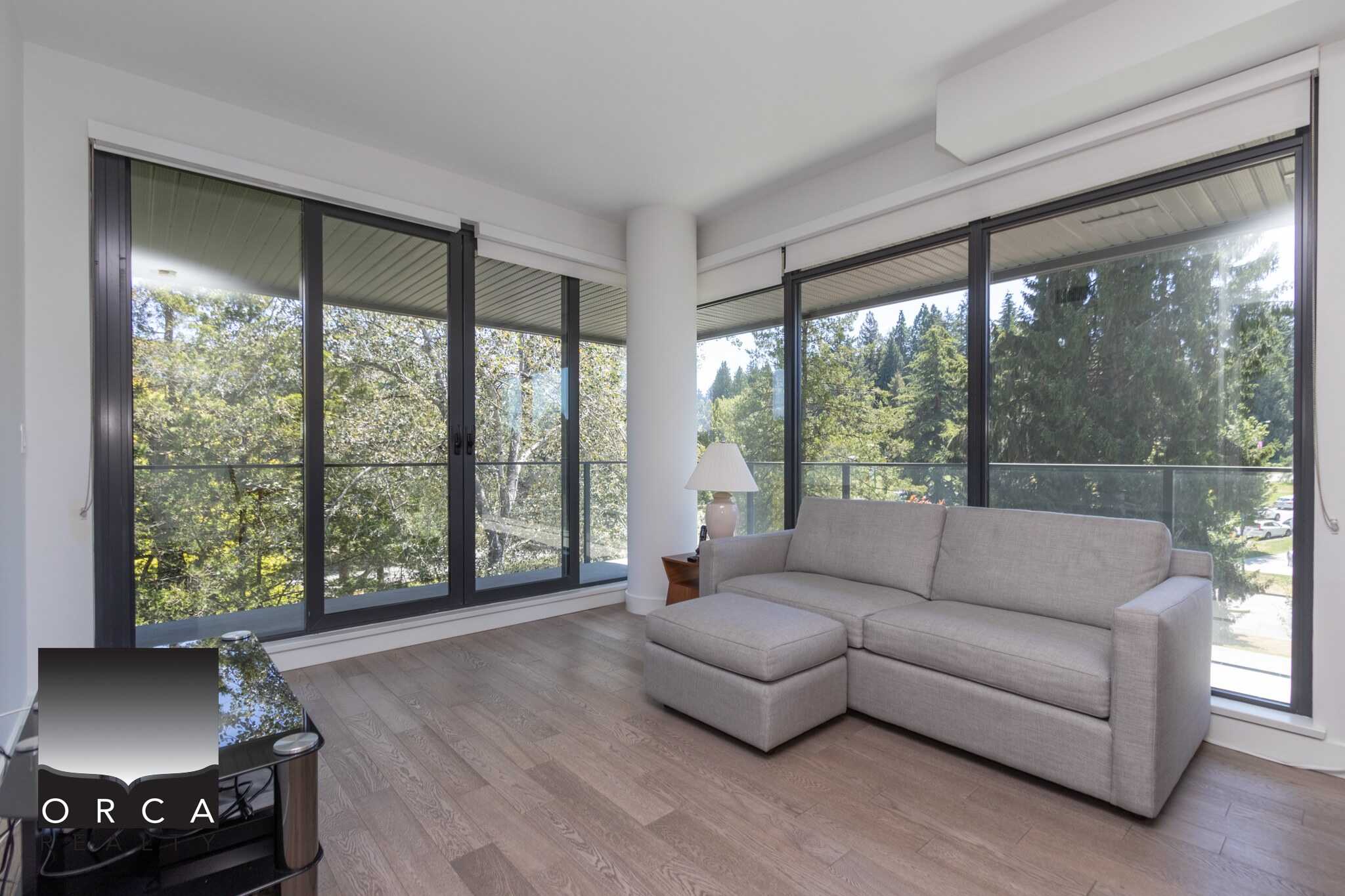 Bright living room with large floor-to-ceiling windows overlooking a lush green trees, featuring a beige sectional sofa, wooden flooring, and natural light, ideal for condo or apartment living in Vancouver.