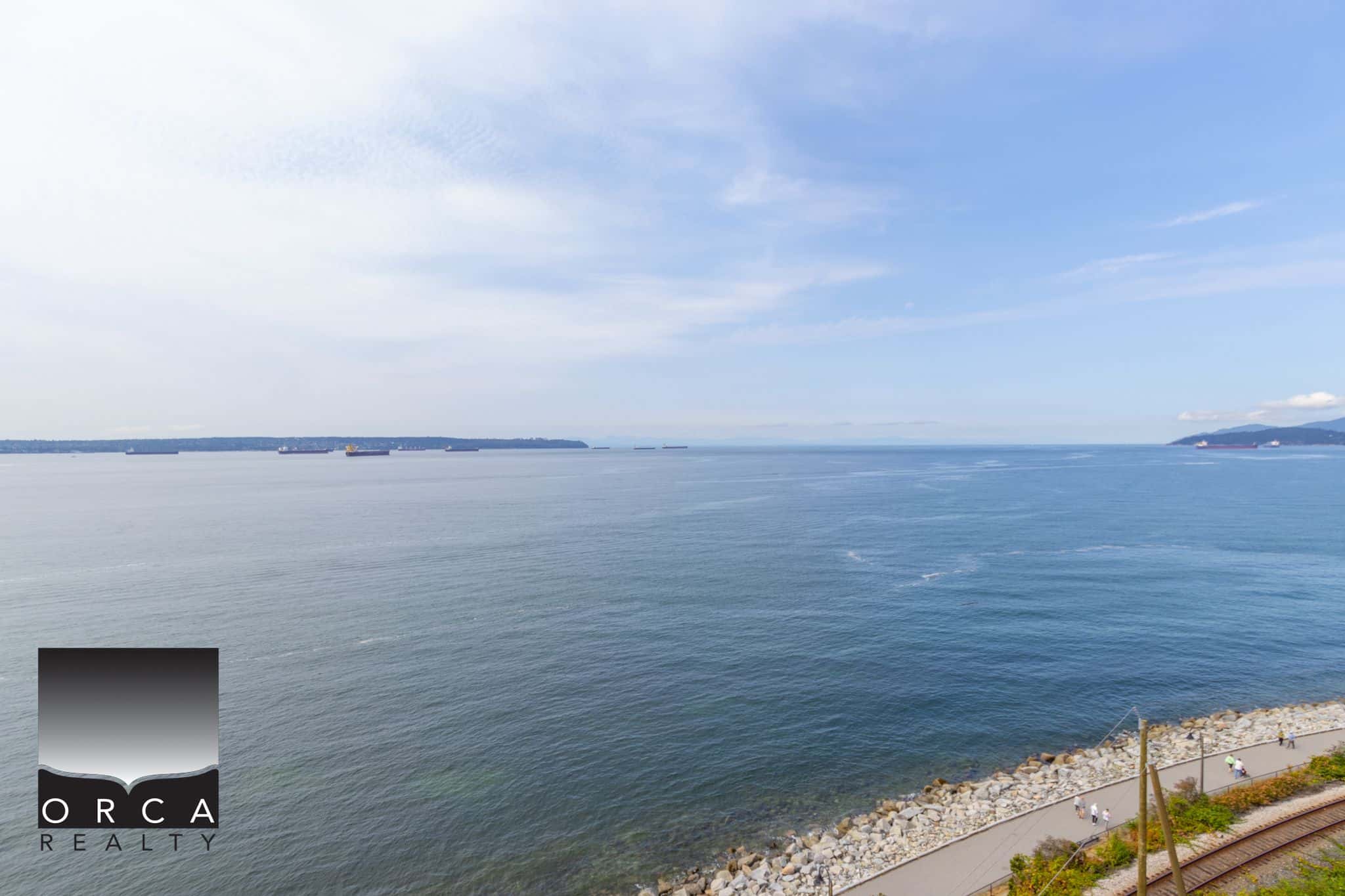 Stunning waterfront view of the ocean with ships sailing, rocky shoreline, and clear blue skies, showcasing the Pacific coastline near Orca Realty Inc. in British Columbia.