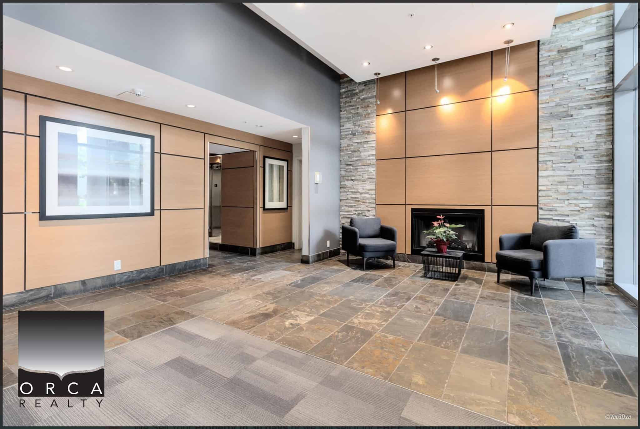 Modern interior lobby with stone and wood accents, two grey armchairs, a glass coffee table with a plant, and a sleek fireplace, showcasing upscale real estate features.