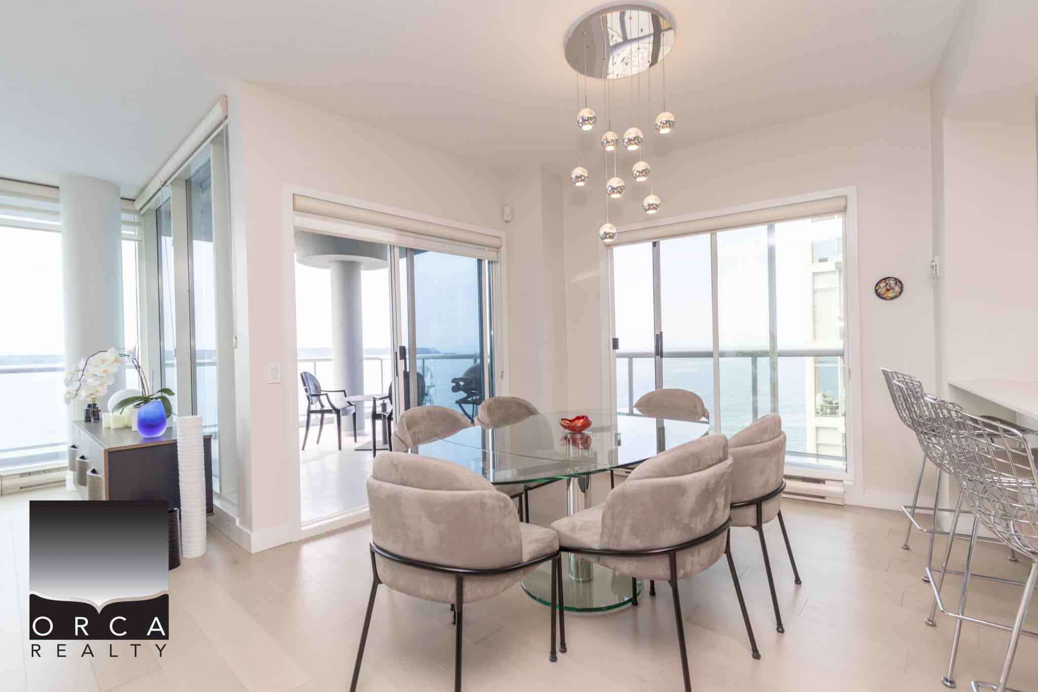 Stylish dining area with ocean views in a modern Vancouver condo, featuring a glass table, plush chairs, and ample natural light from large windows and balcony access.